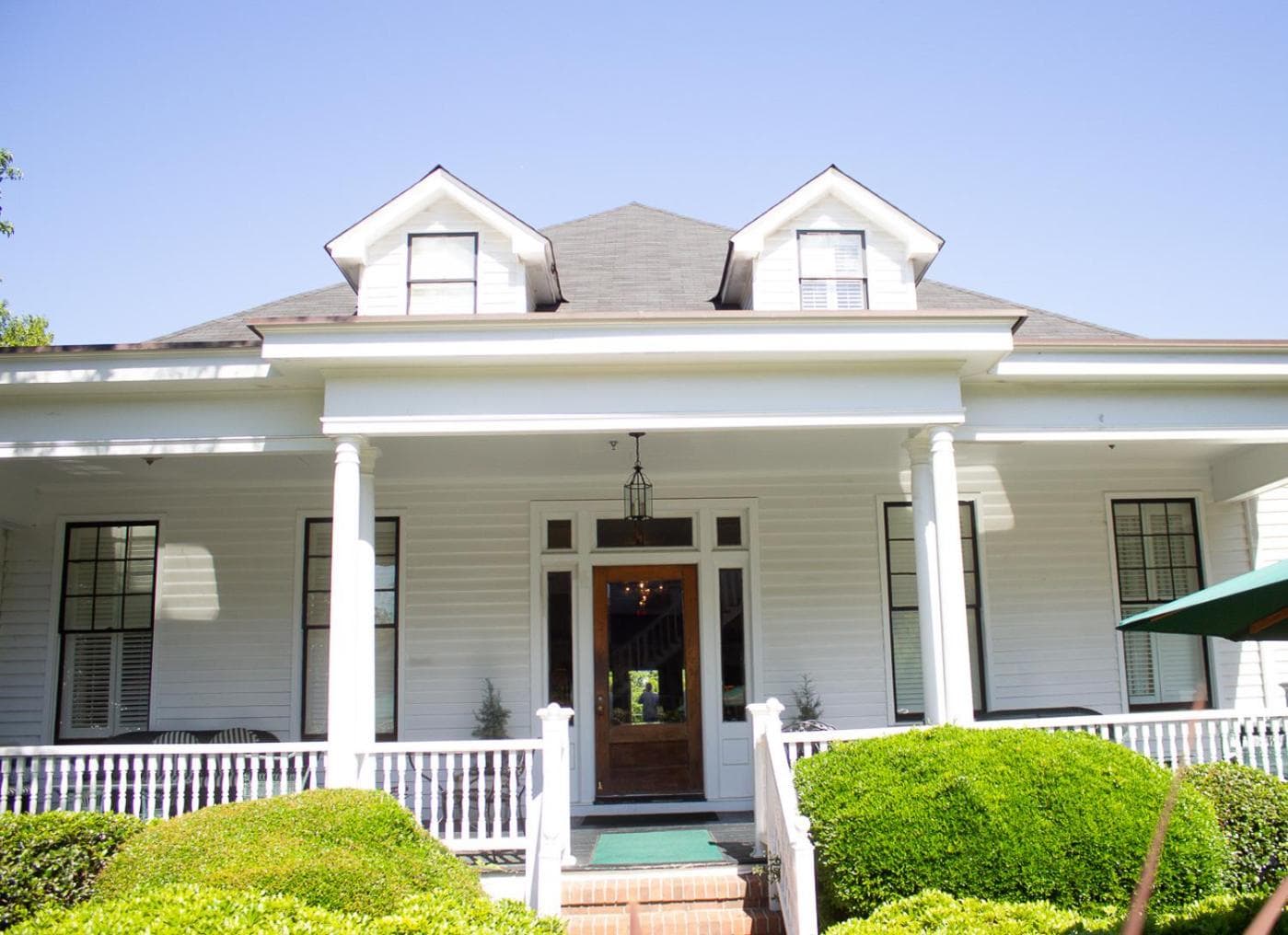 White cottage facade with a covered porch, railing, trimmed boxwoods, and dormer windows on the roof.