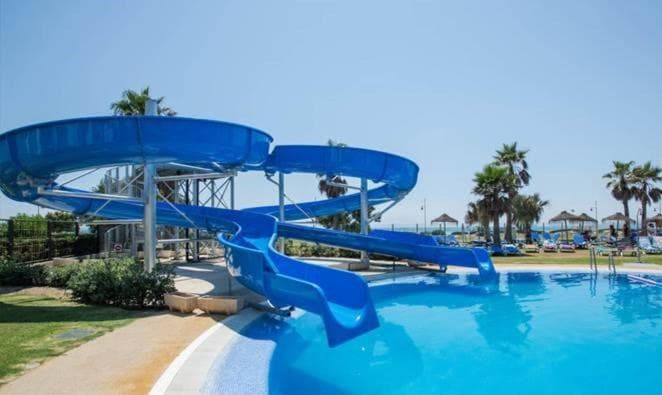 A blue spiral water slide curves into an outdoor swimming pool bordered by palm trees.