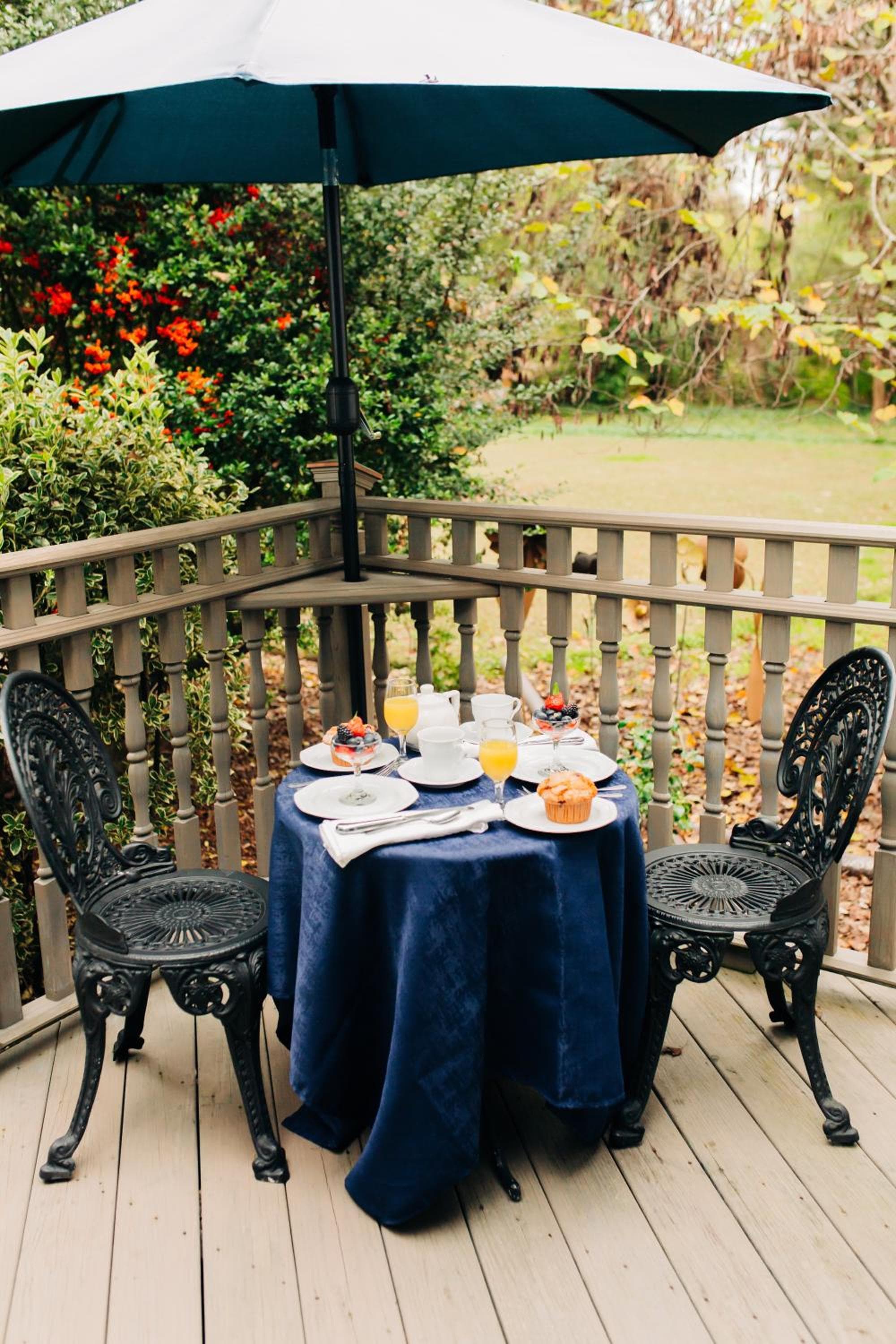 An outdoor deck table covered in dark cloth holds breakfast food and drinks beside wrought iron chairs.
