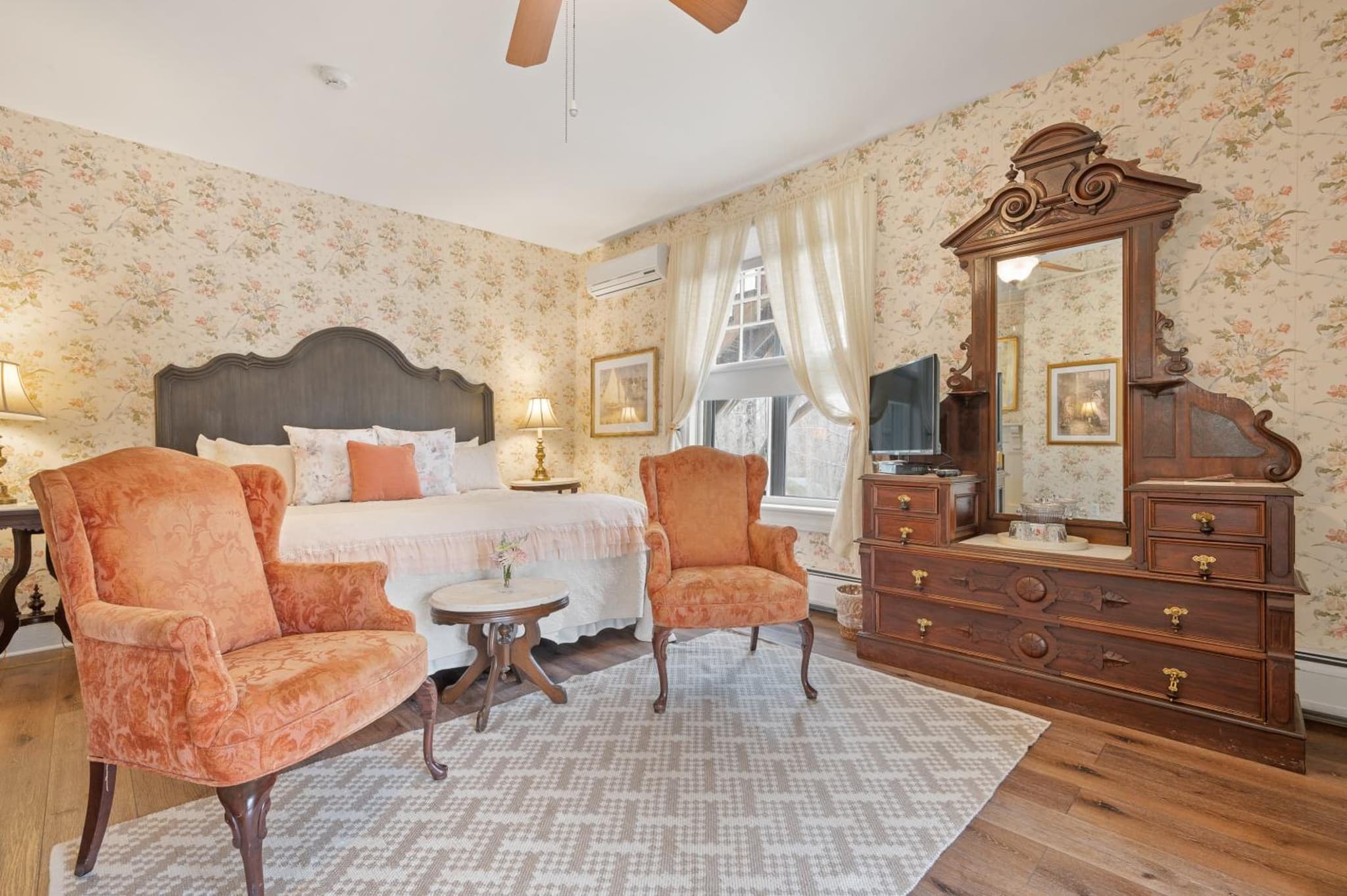 A guest room with floral wallpaper, an ornate carved wooden dresser and mirror, upholstered armchairs, and a bed.