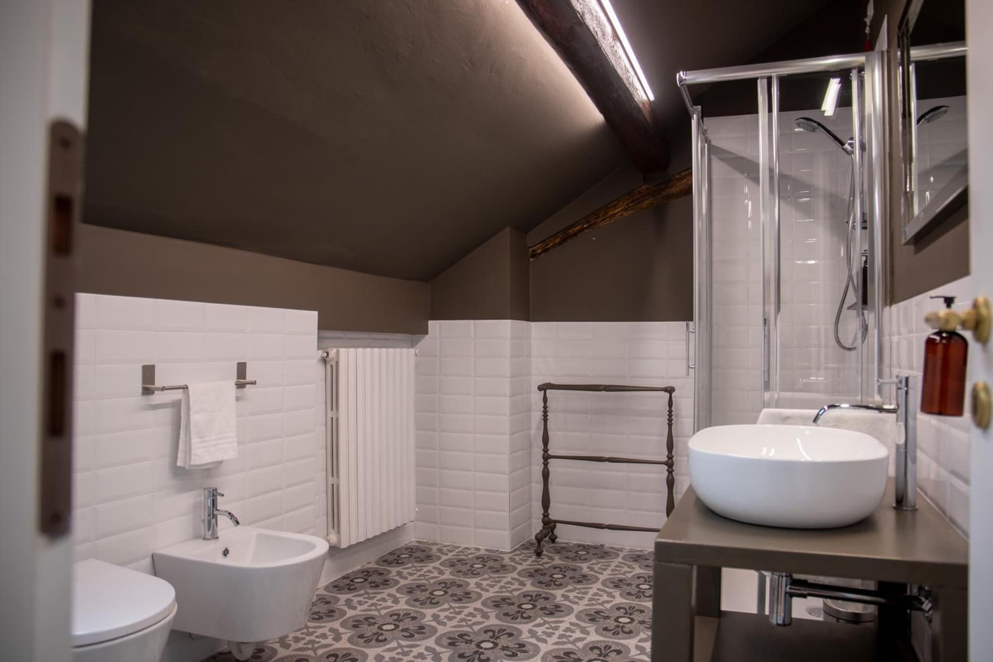 An attic bathroom with patterned floor tiles, white subway walls, an exposed ceiling beam, and a vessel sink.
