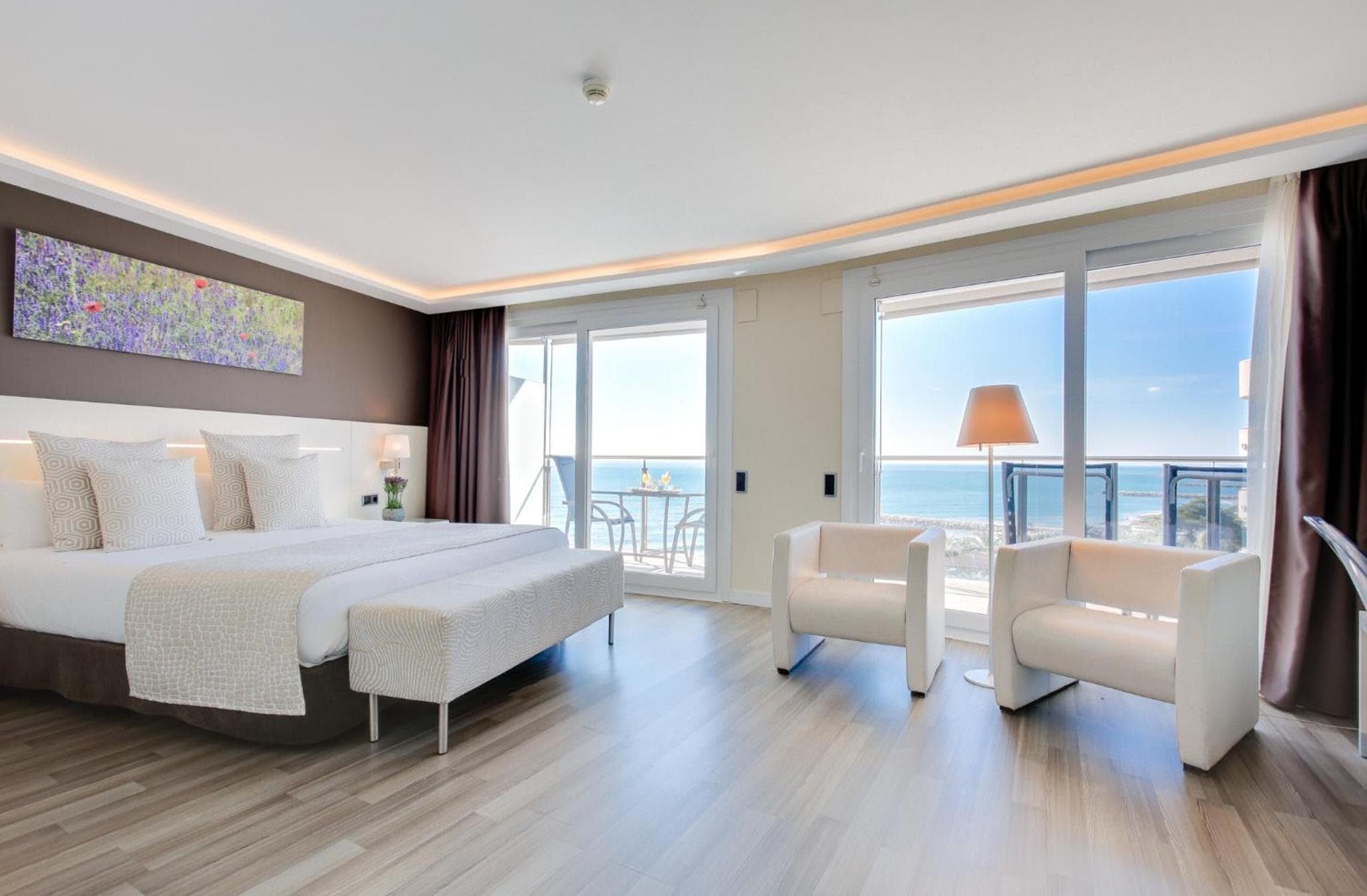 Bedroom with wood-look flooring, white armchairs, and sliding glass doors opening to a sea view.