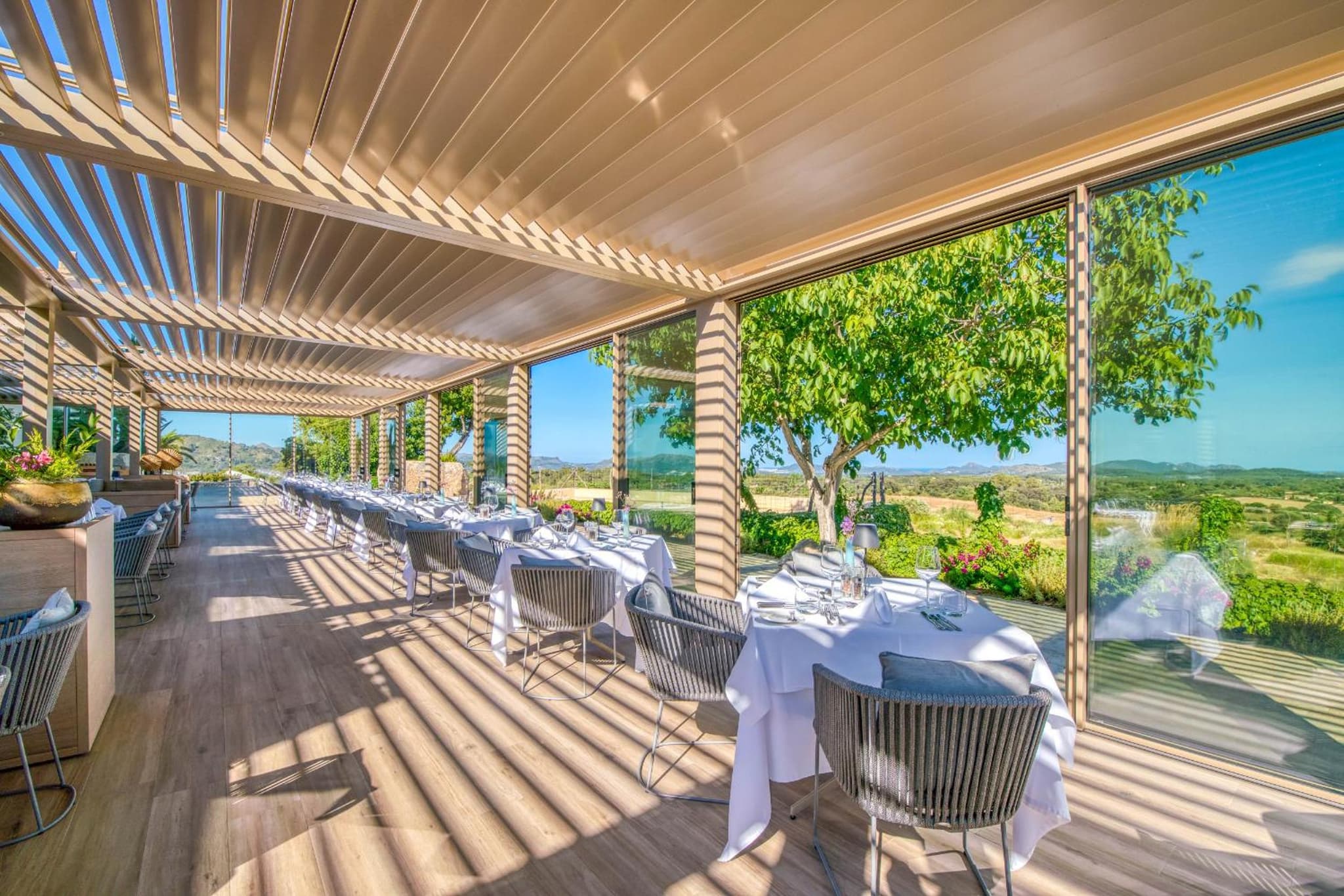 A covered terrace with slatted pergola, set dining tables, and open views over the countryside.