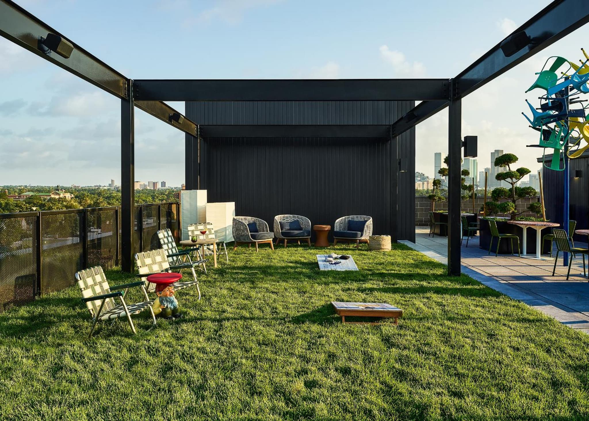 A rooftop terrace with a green lawn under a dark steel pergola, furnished with patio seating and lawn games.