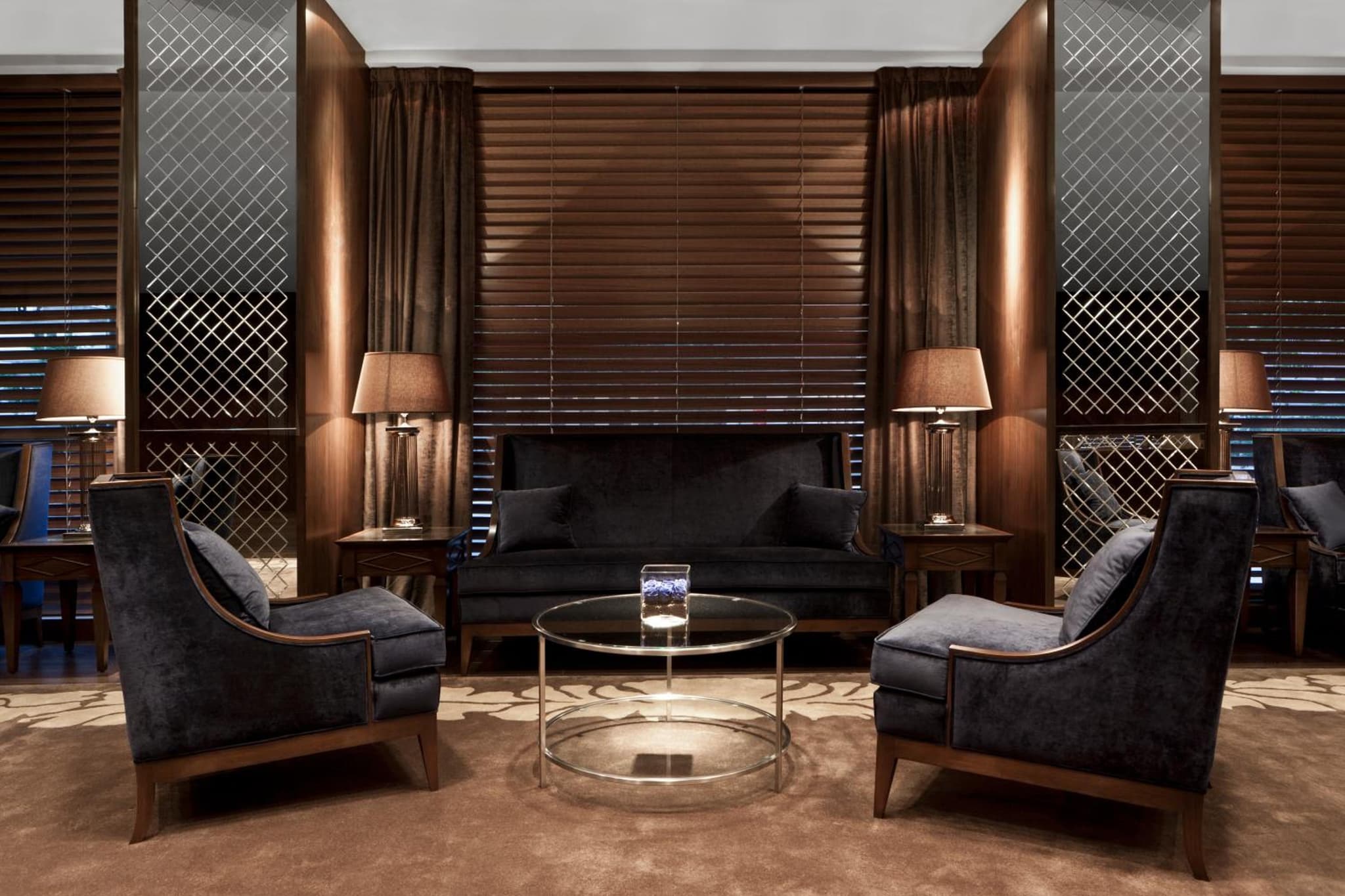 Indoor lounge with dark upholstered armchairs, glass coffee table, warm lamps, and dark wood wall panels.