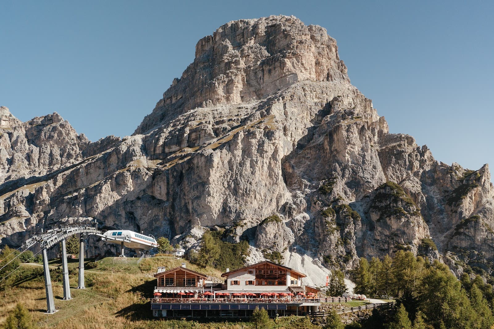 A timber and render mountain refuge with a red-umbrella terrace sits below a sheer rocky Dolomite peak, with a cable car visible to the left.