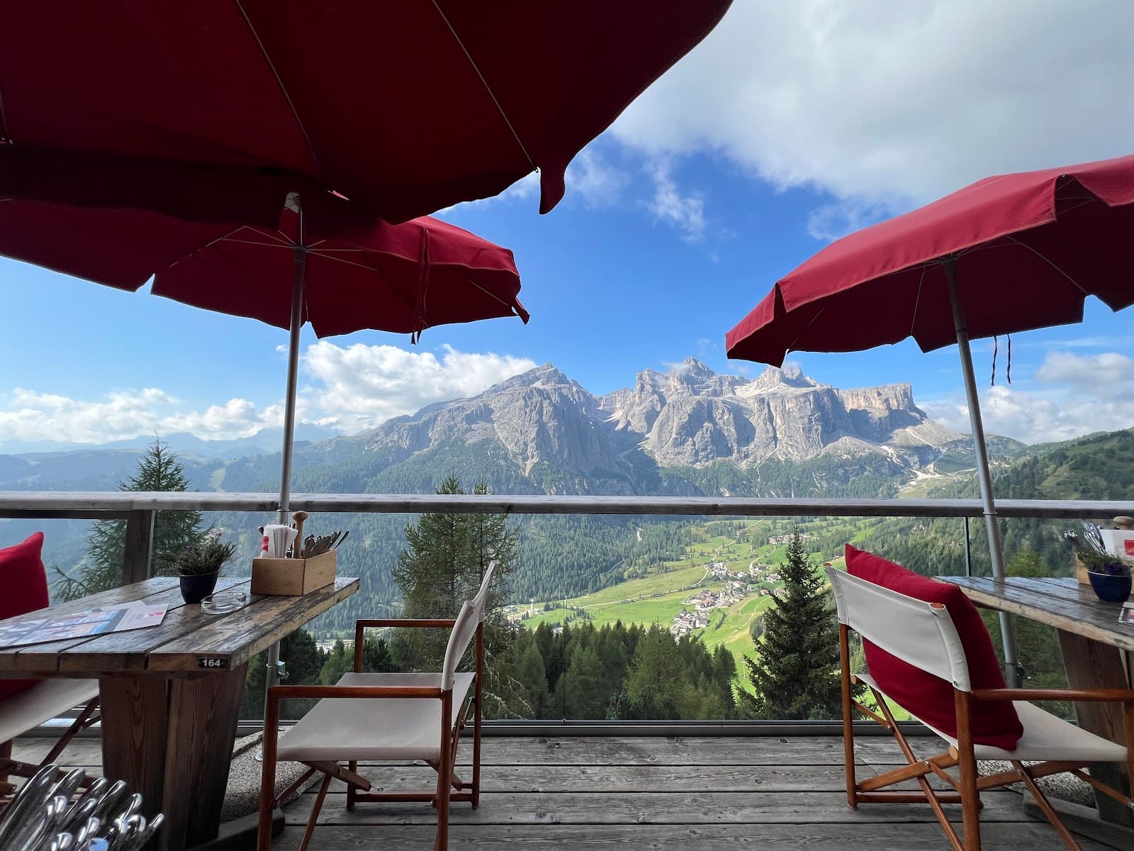 A timber deck terrace with red umbrellas and canvas chairs overlooking a wide valley of forest and meadows backed by pale Dolomite rock faces under a blue sky.