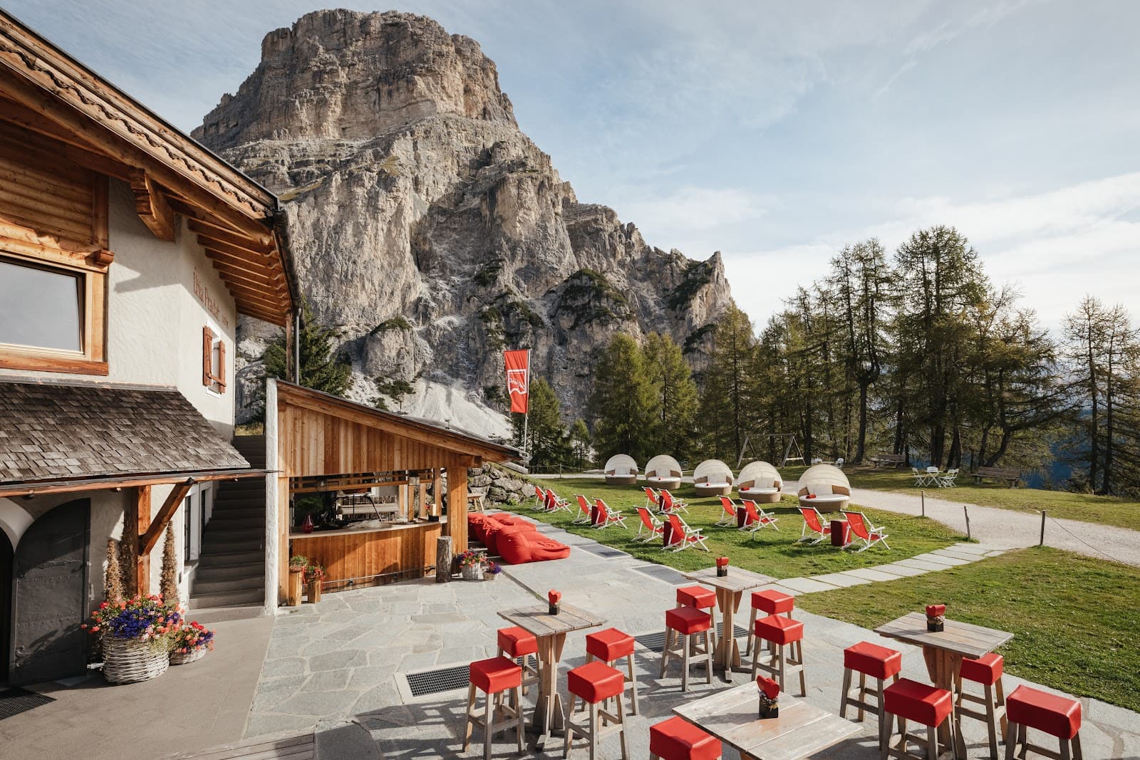 The stone-paved outdoor bar terrace of Col Pradat with red bar stools, a wooden counter, red sun loungers on a grassy lawn, and a rocky cliff face behind.