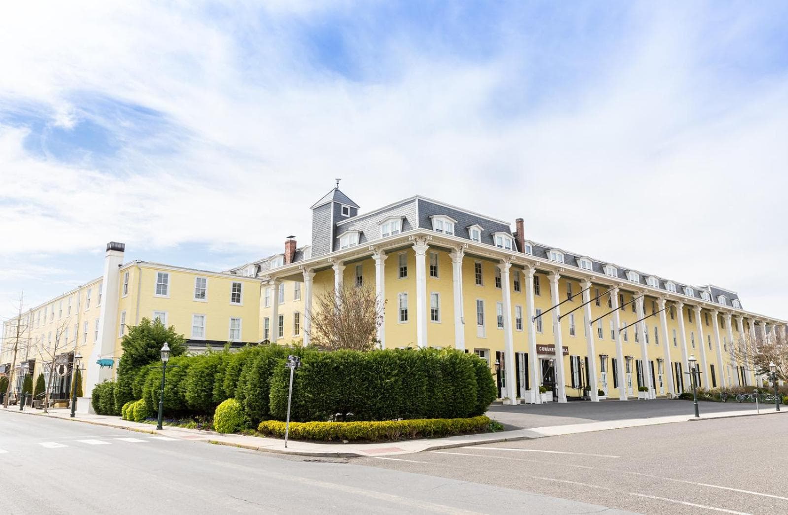 The exterior of the large yellow hotel bordered by manicured hedges under a partly cloudy sky.