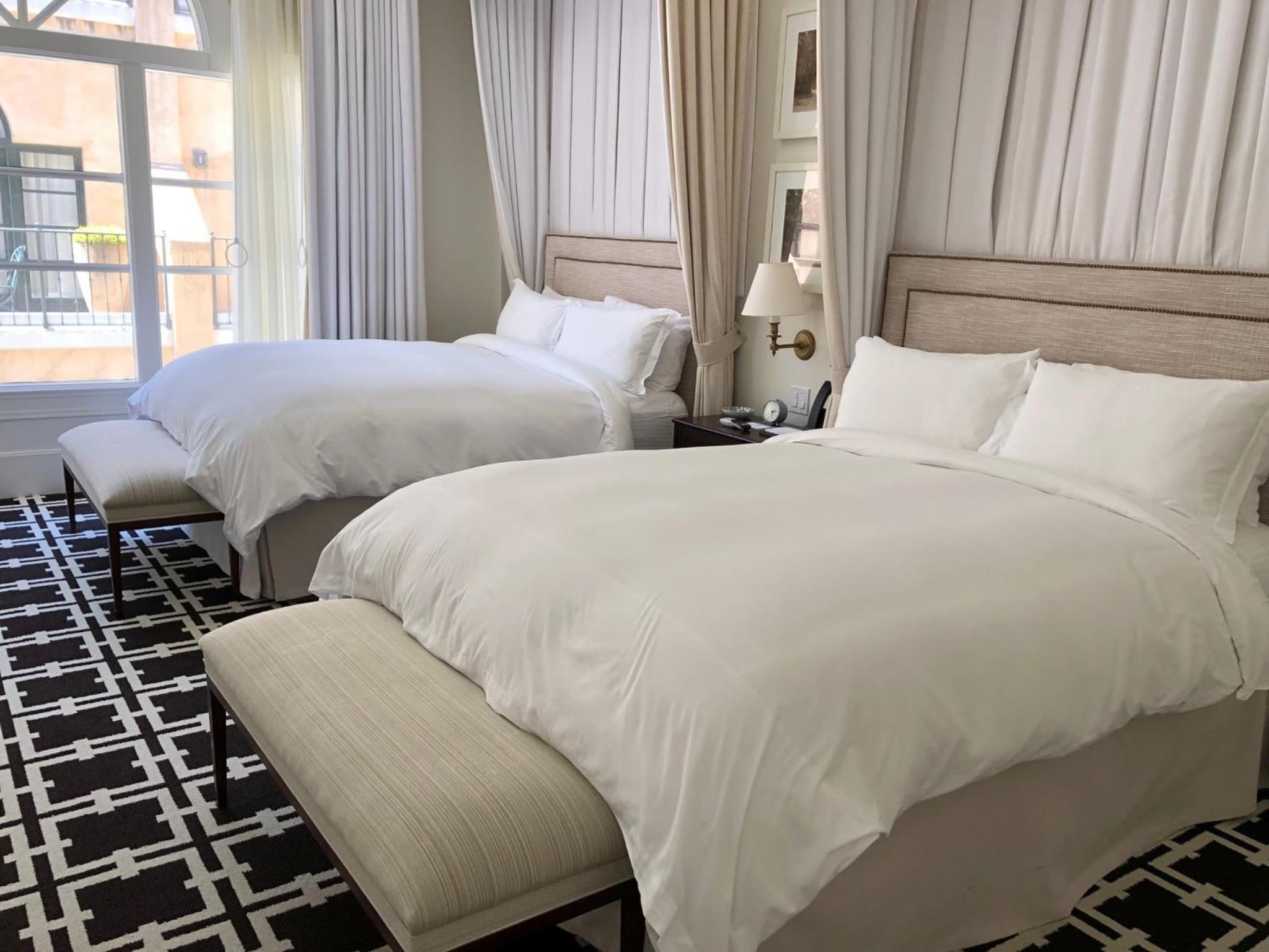 Guest room with beds dressed in white linens, upholstered headboards, a patterned rug, and sheer drapes by a terrace door.