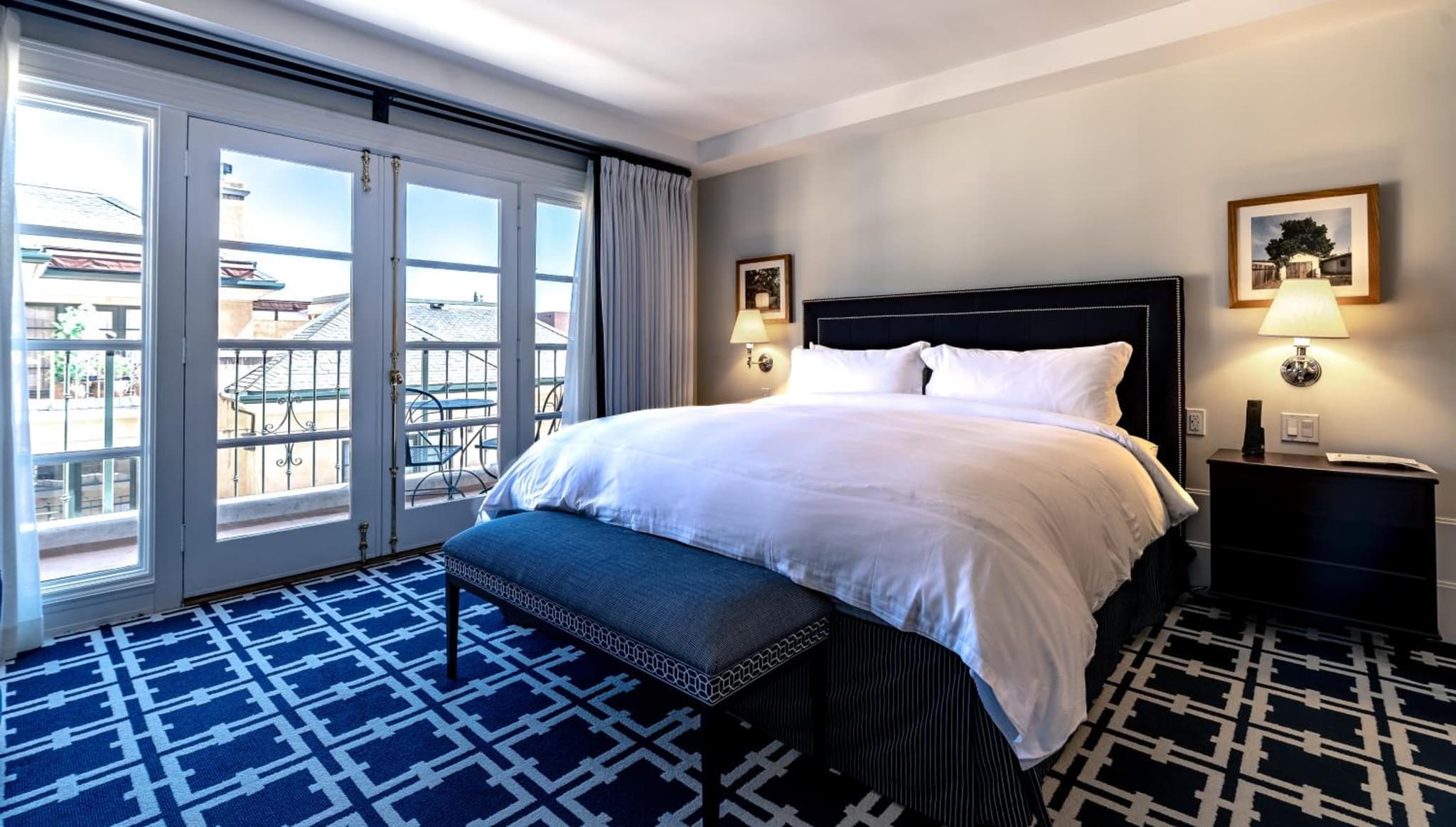 Bedroom with a dark cushioned headboard, white bedding, geometric blue carpet, and French doors opening onto a balcony.