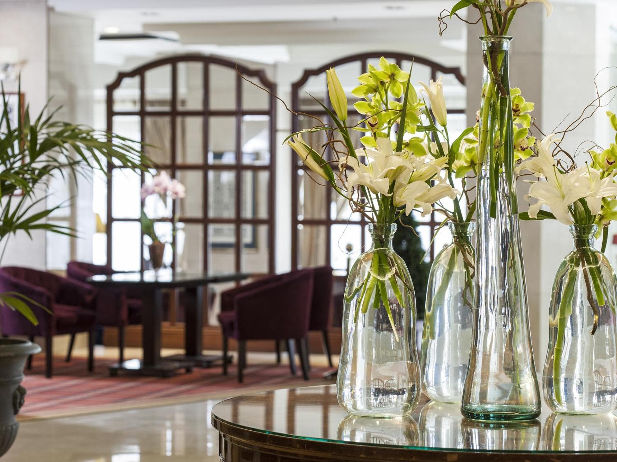 Tall glass vases with white and green flowers set atop a round glass table in a lounge area.
