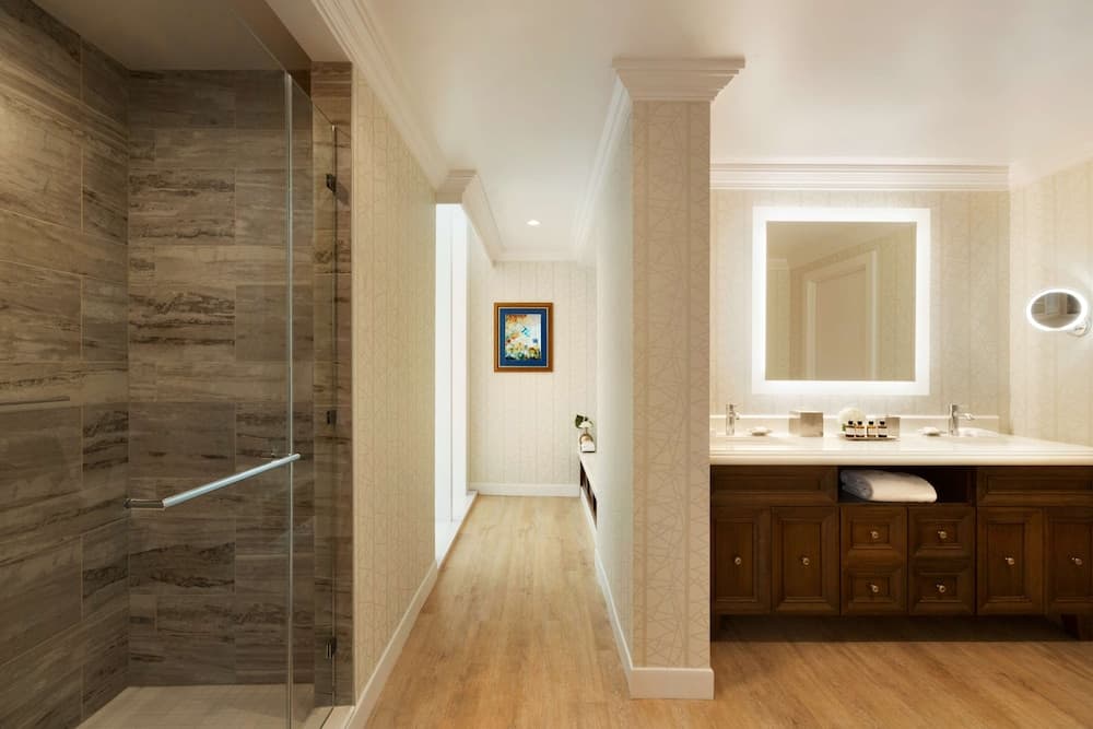 A hotel bathroom featuring a glass shower stall, wood vanity with a backlit square mirror, and light flooring.