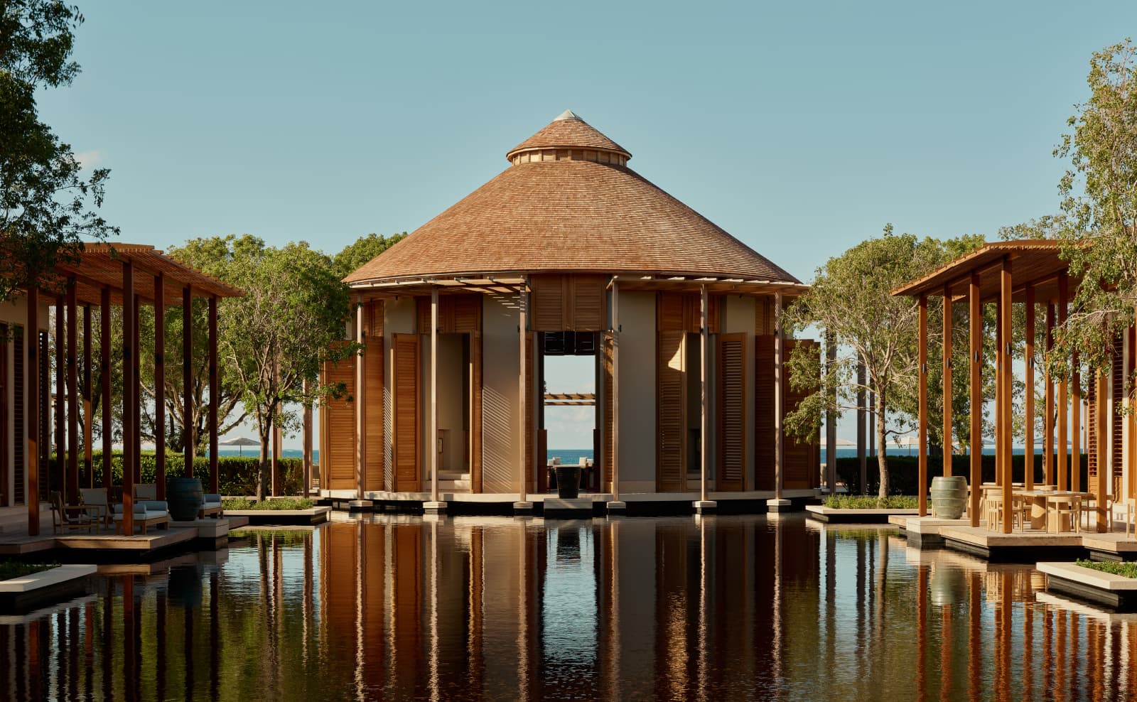 A circular pavilion with a conical timber roof reflects across a still pool flanked by pergolas and trees.