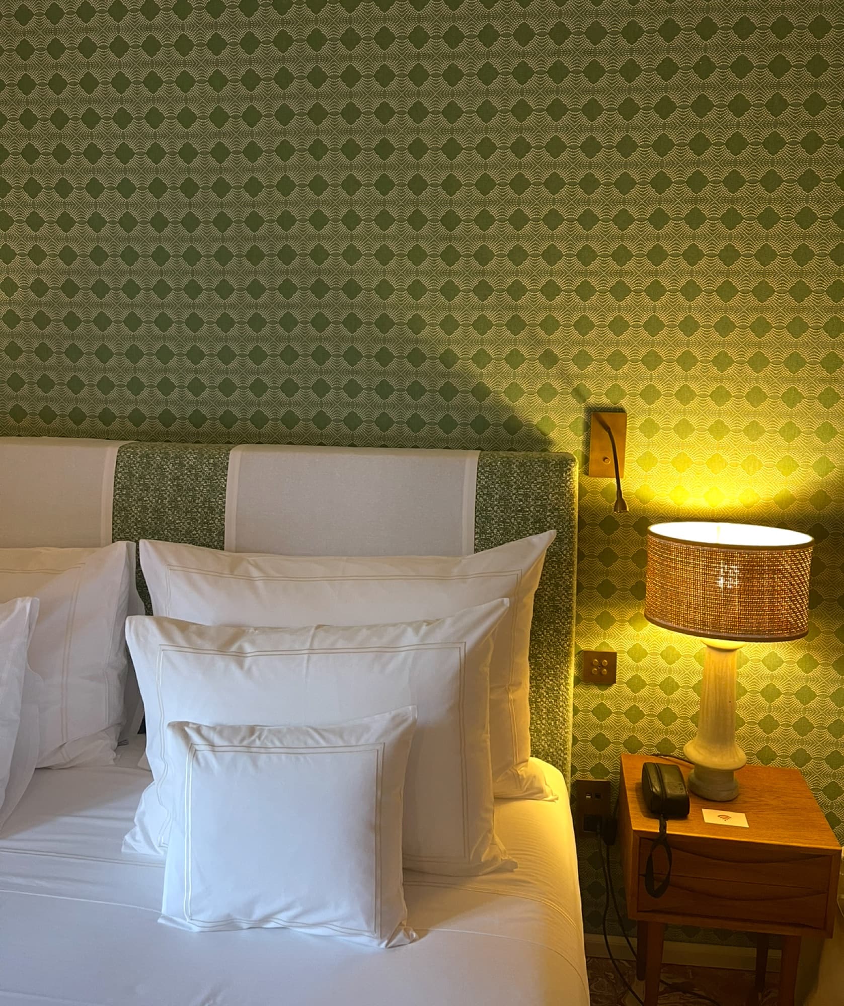 White bed linens rest beside a wooden nightstand with an illuminated woven lamp against green patterned wallpaper.