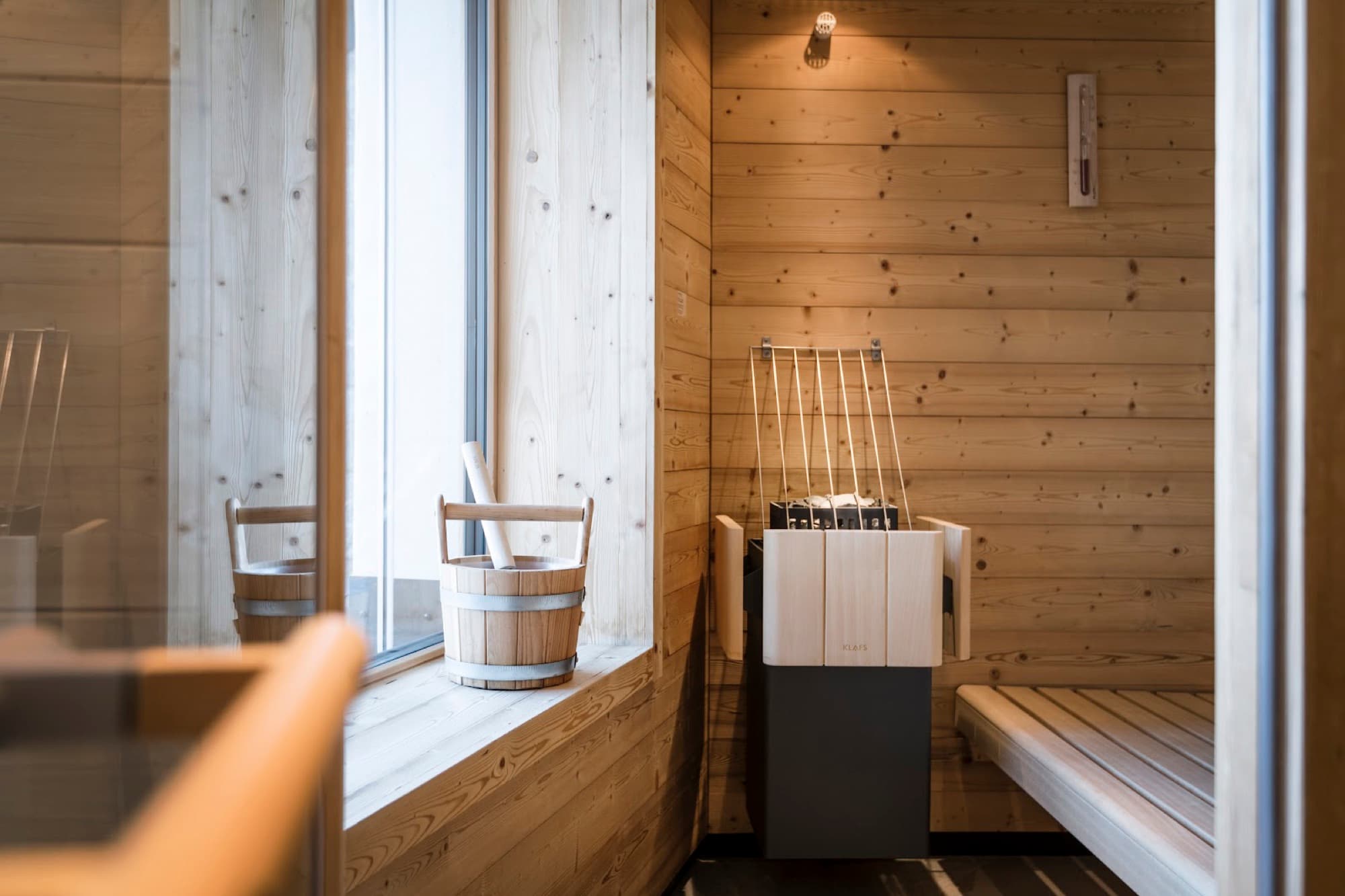 A wooden sauna interior with a heater, bench, and wooden water bucket on a deep windowsill beside a large window.