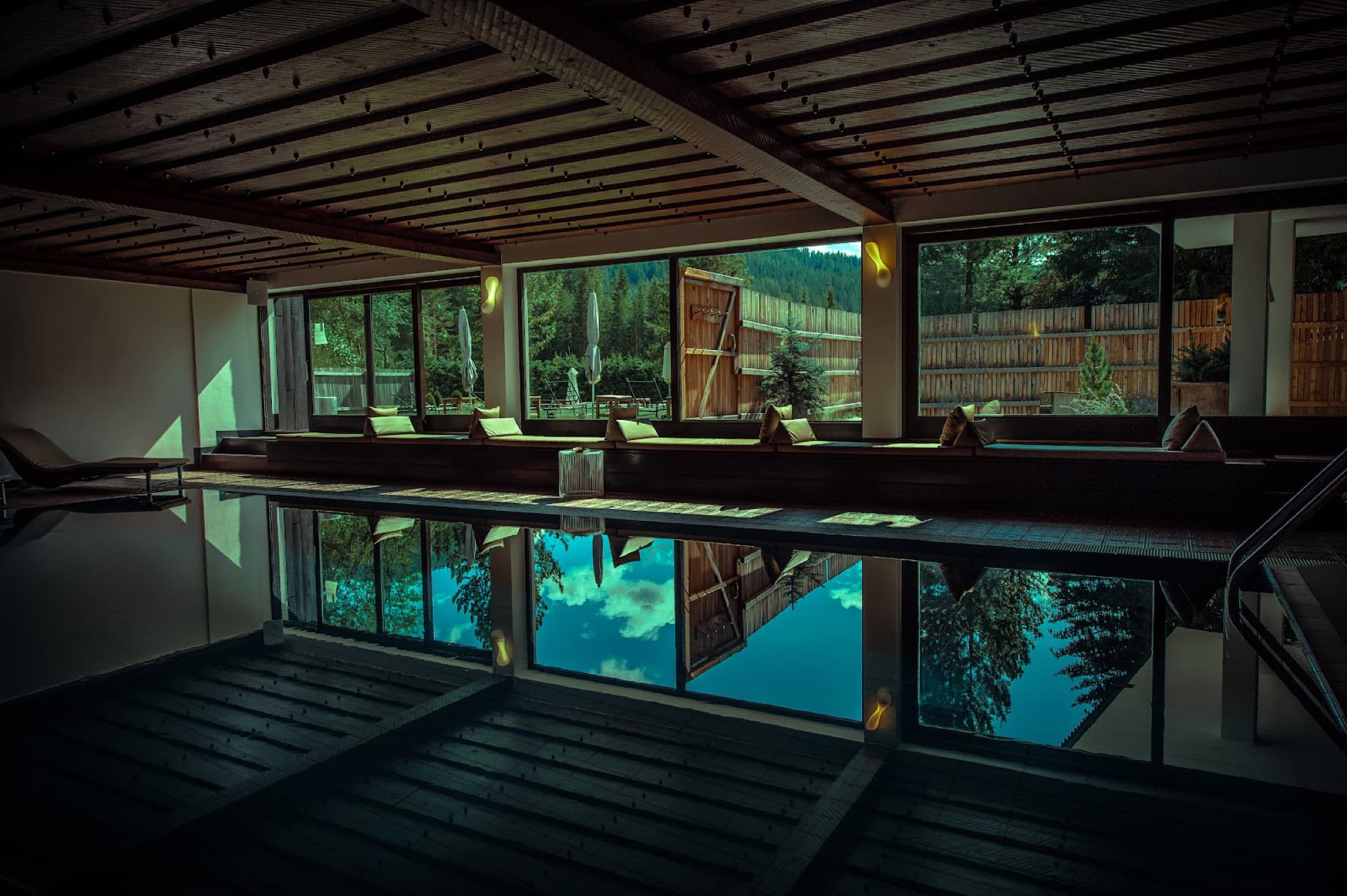 An indoor pool under a dark slatted timber ceiling with large windows reflecting outdoor pine trees and sky.