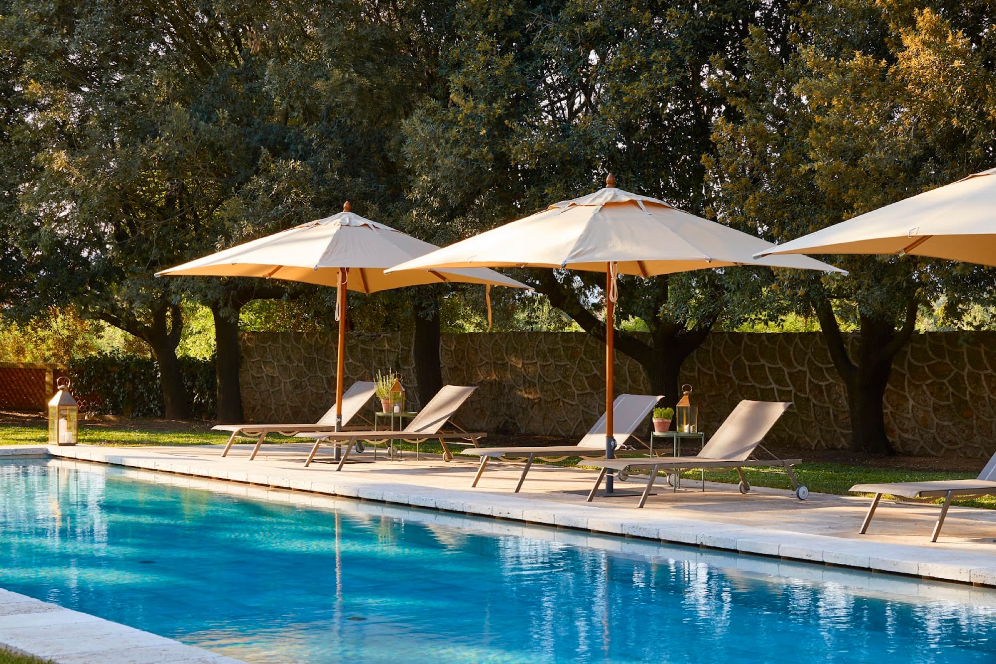 Sun loungers and umbrellas along the edge of a swimming pool backed by stone walls and mature trees.