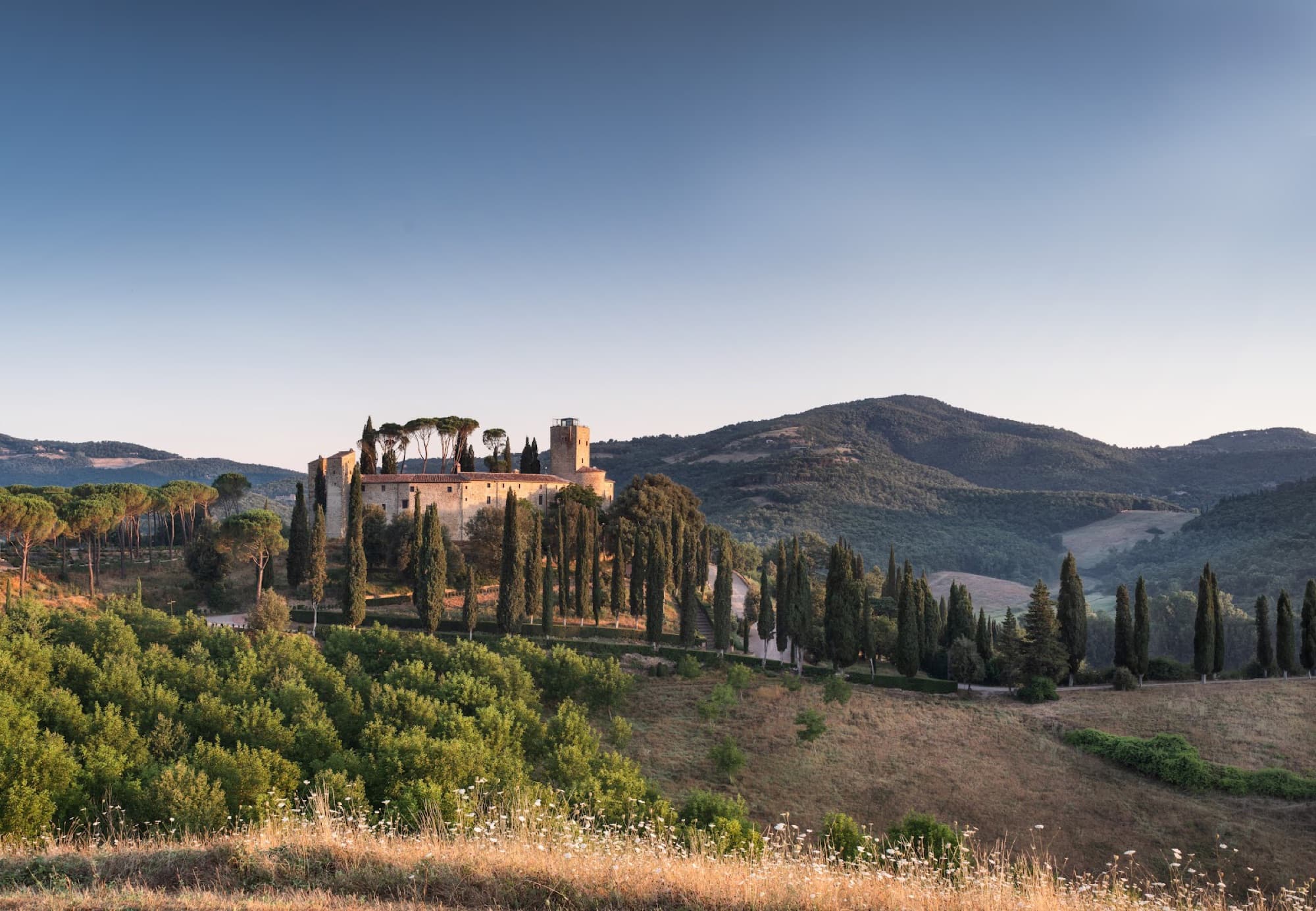 Castello di Reschio stands on a hilltop framed by tall cypress trees across rolling green valleys.