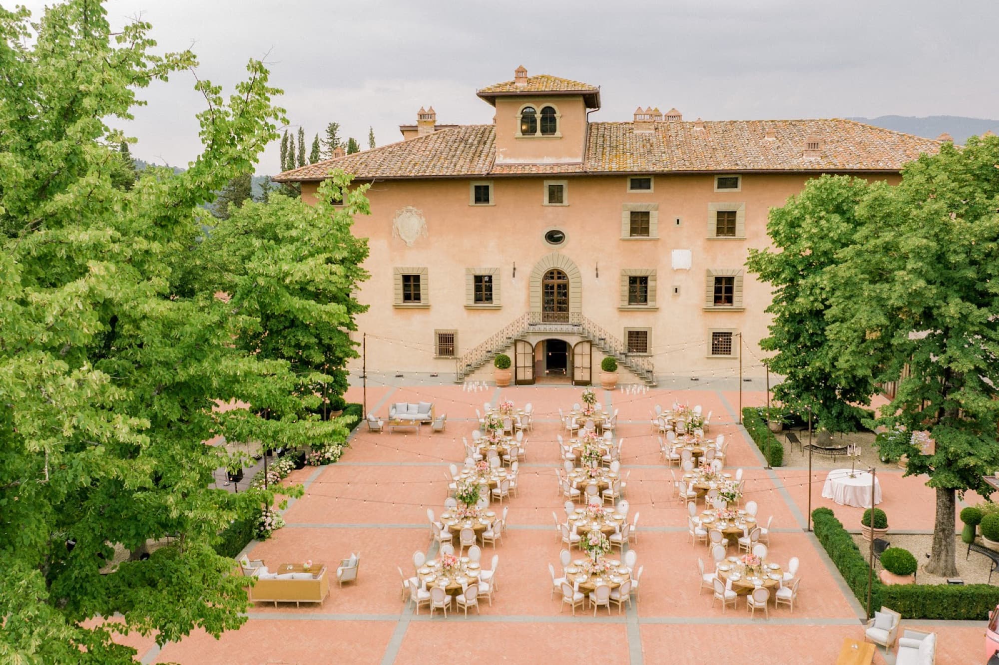 A terracotta courtyard arranged with round banquet tables and floral centerpieces before the main villa.