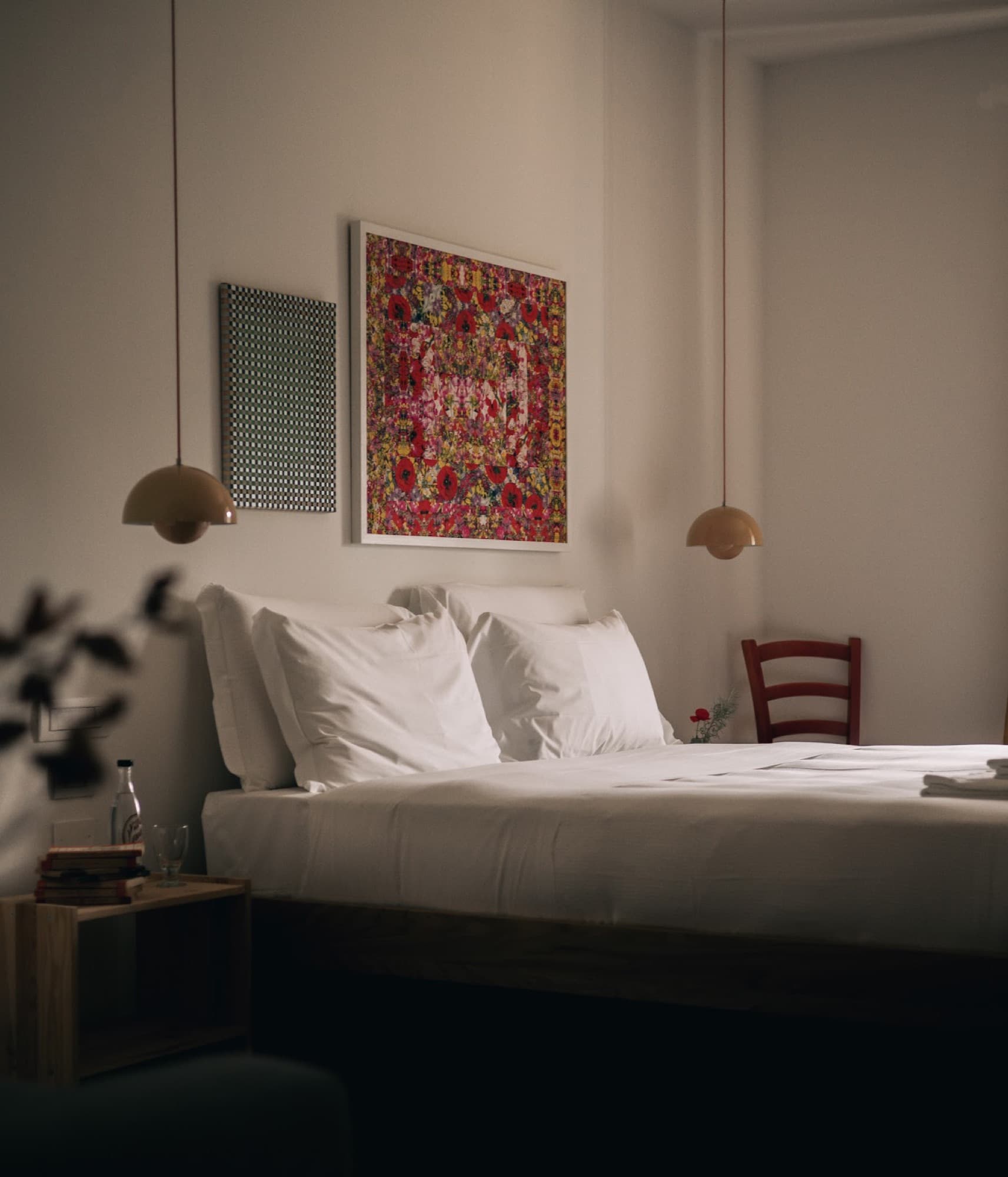 A bedroom with white bedding, hanging pendant lights, and colorful artwork on the white wall.