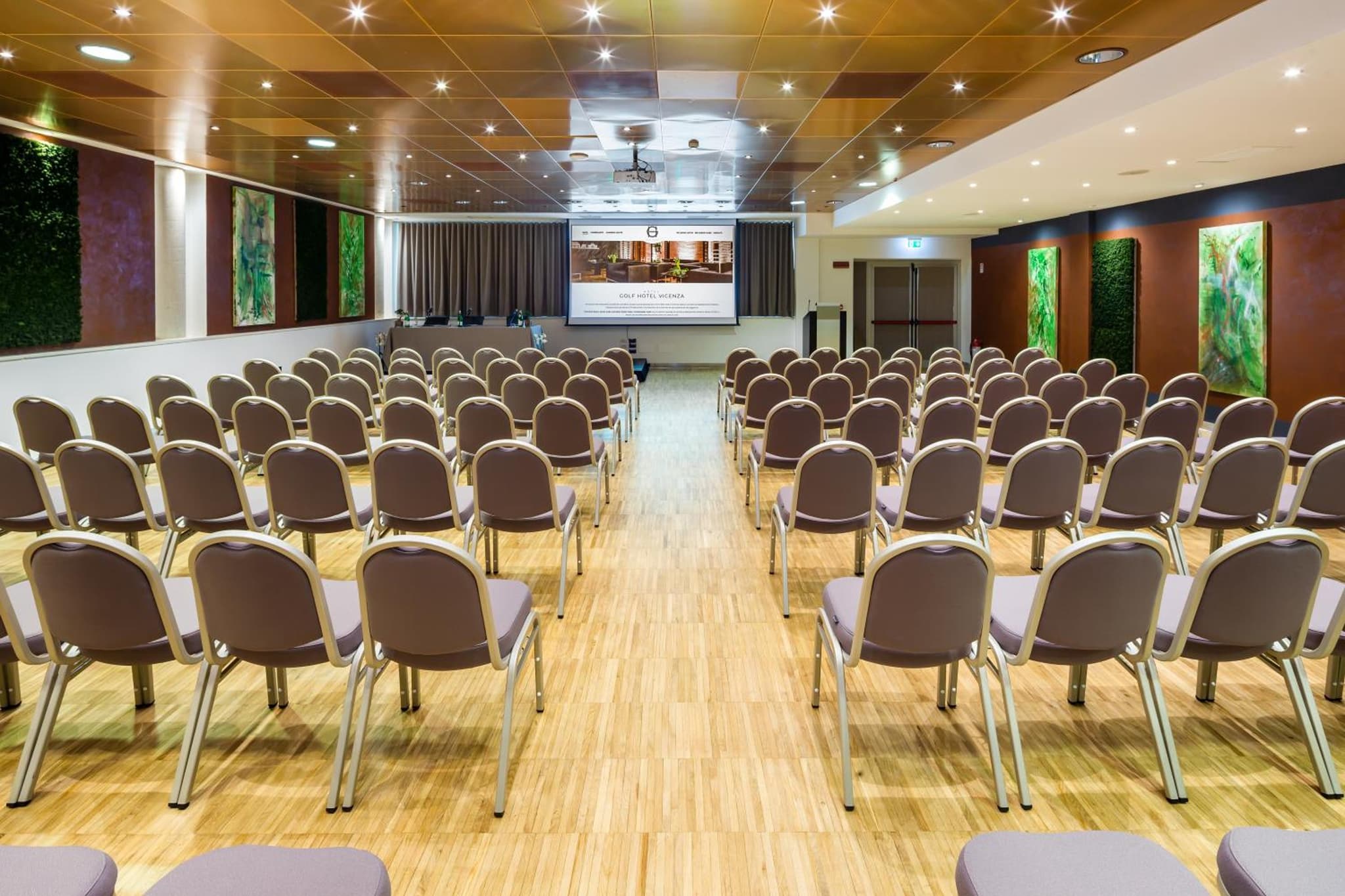 Conference hall with rows of upholstered chairs facing a projection screen over parquet flooring.