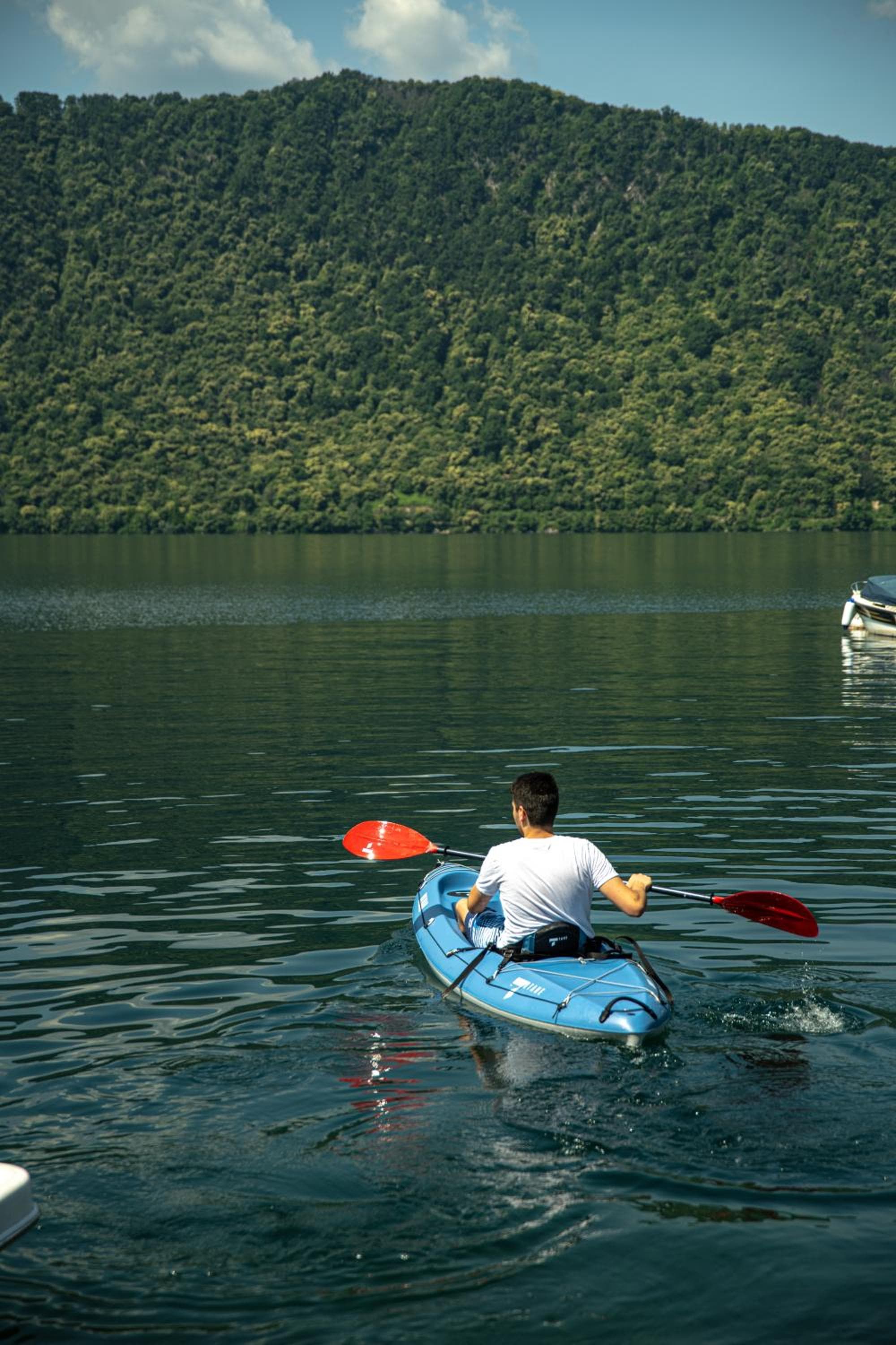 A kayaker paddling across calm lake water toward a steep, tree-covered mountain.