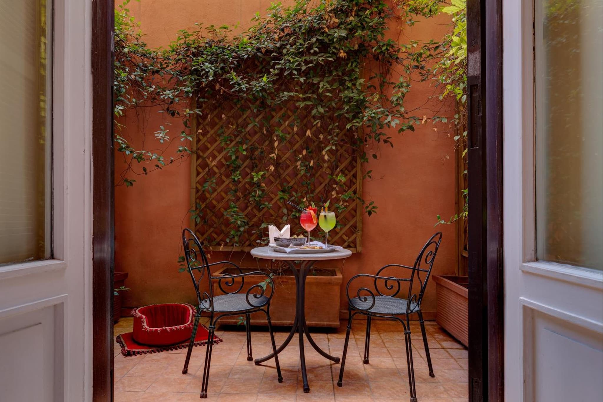 An interior courtyard patio with terracotta walls, climbing vines, and metal chairs beside a drink table.
