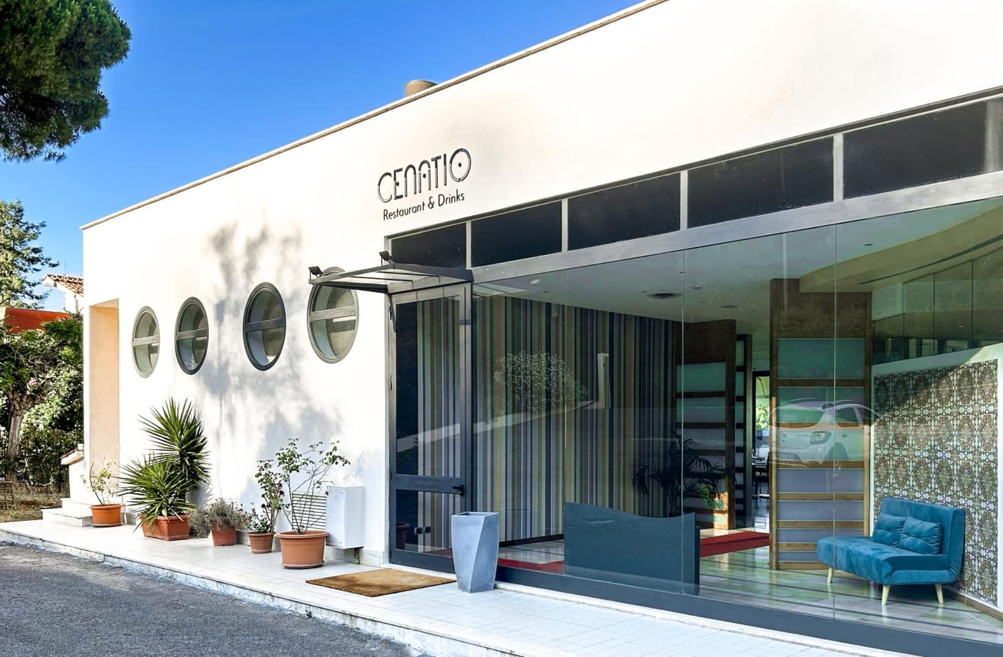 Bright exterior view of Cenatio restaurant with expansive glass windows and potted plants on a white terrace.