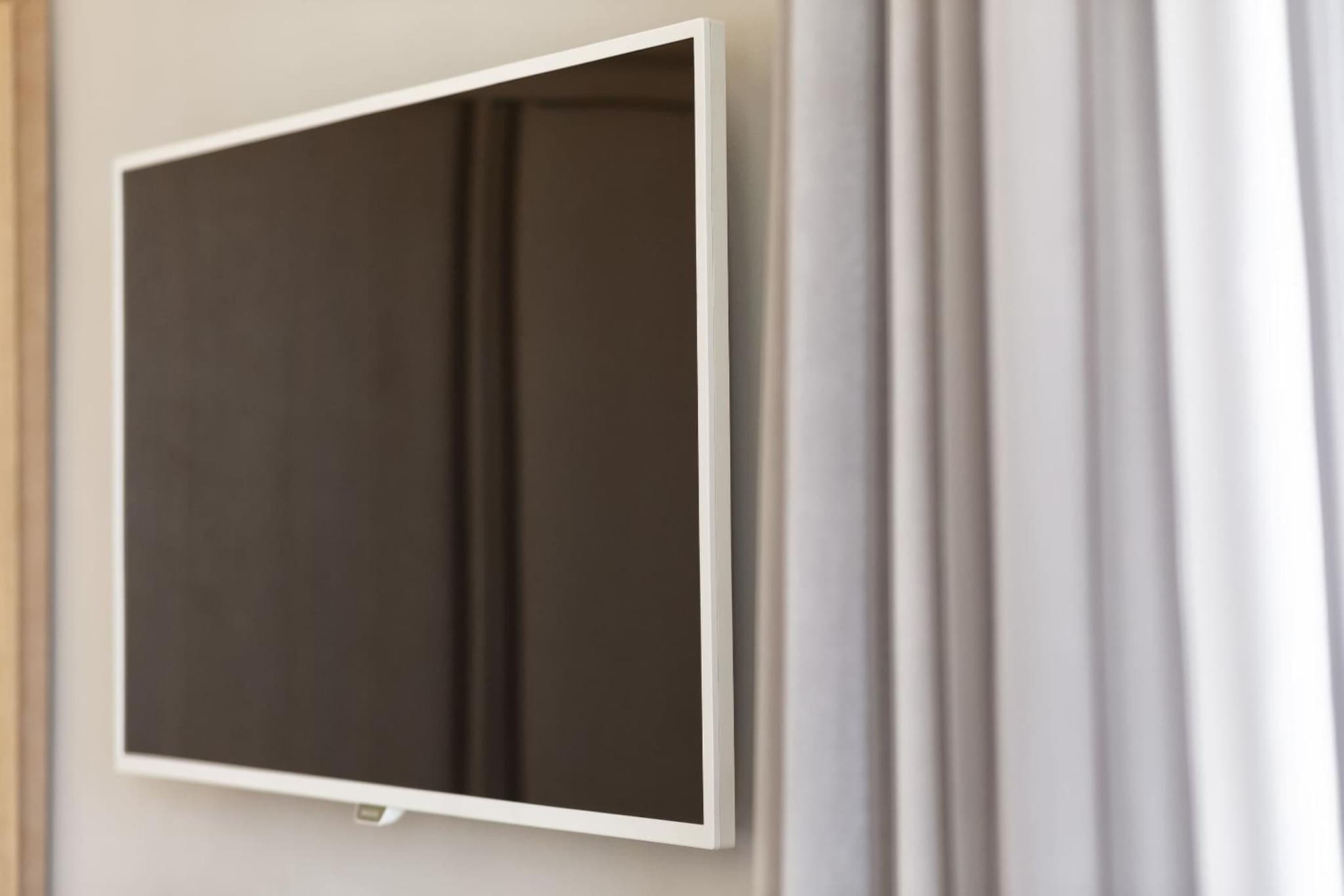 A flat-screen television mounted on a neutral wall beside gathered sheer curtains.