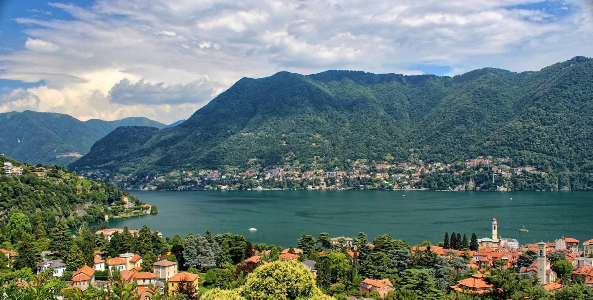 High-angle view across Lake Como toward forested mountains, lakeside towns, and a church tower.