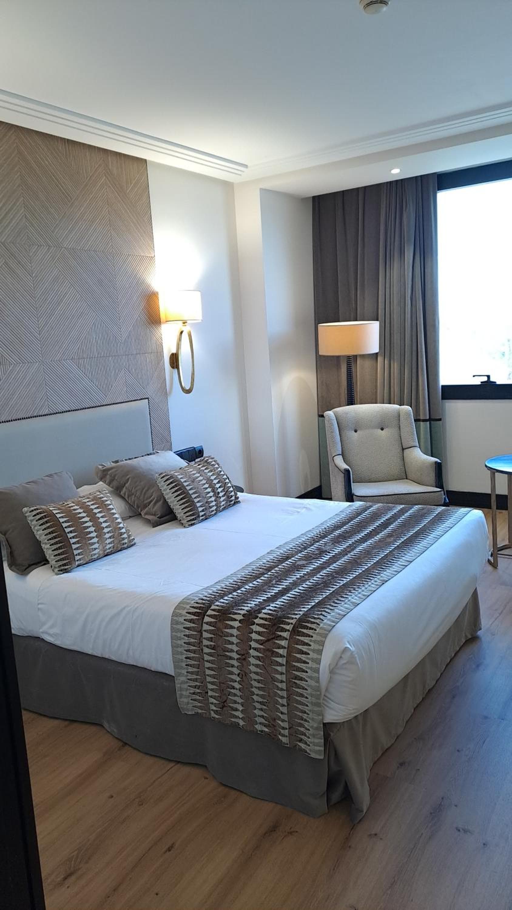 A made bed with patterned accent pillows and runner, beside an upholstered armchair and draped window.