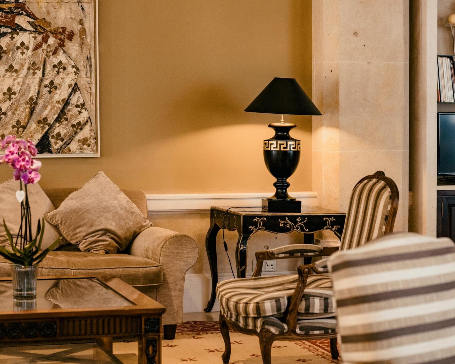 A lounge corner with a striped armchair, a velvet sofa, an orchid, and a classical black urn table lamp.
