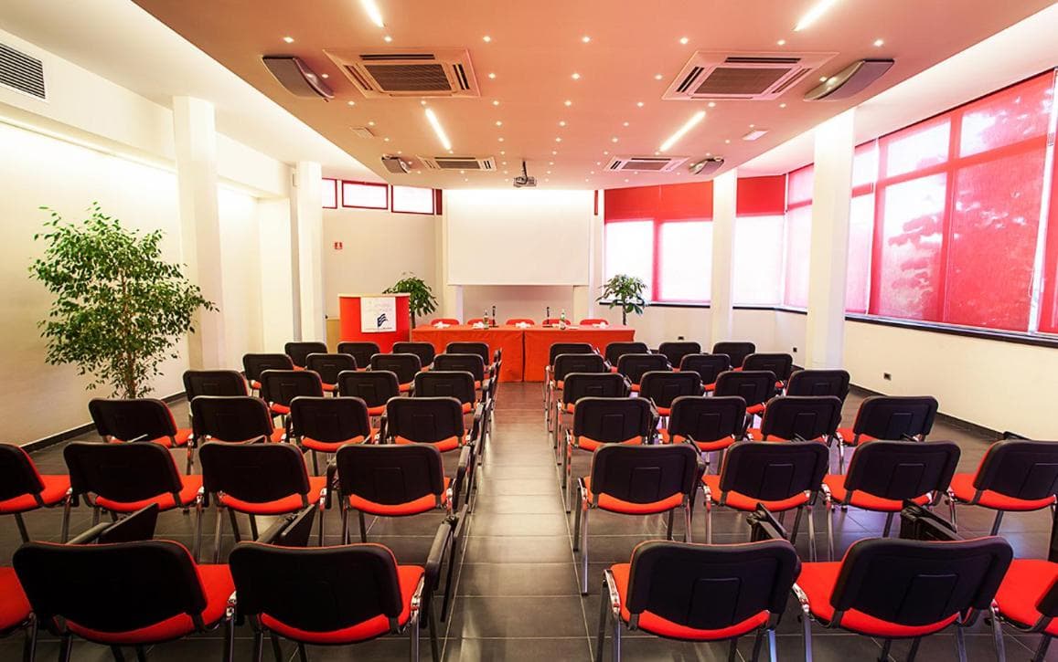 Meeting room arranged with rows of black and red chairs facing a presentation table and projector screen.