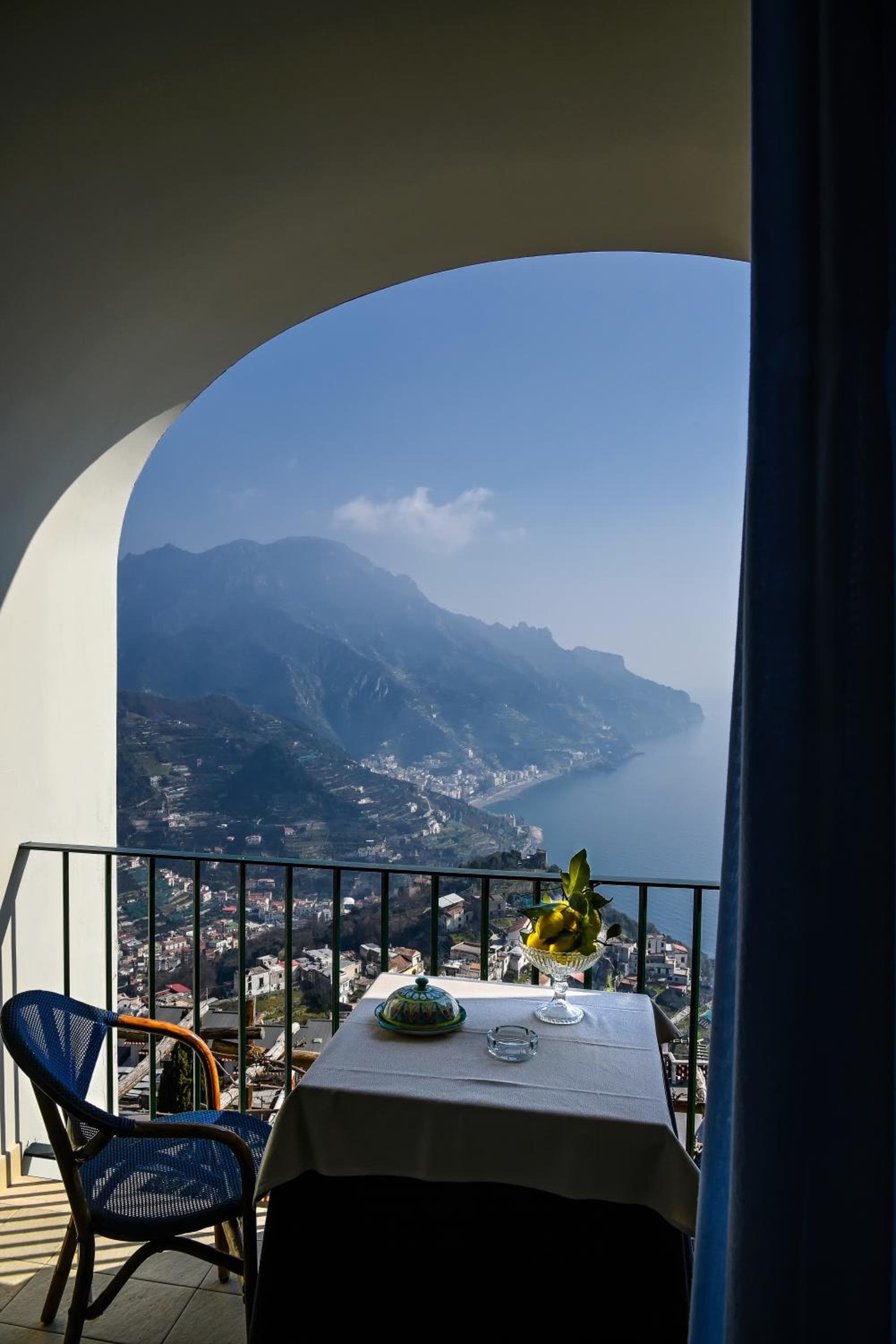 An arched opening frames a balcony table with a bowl of lemons overlooking the coast.