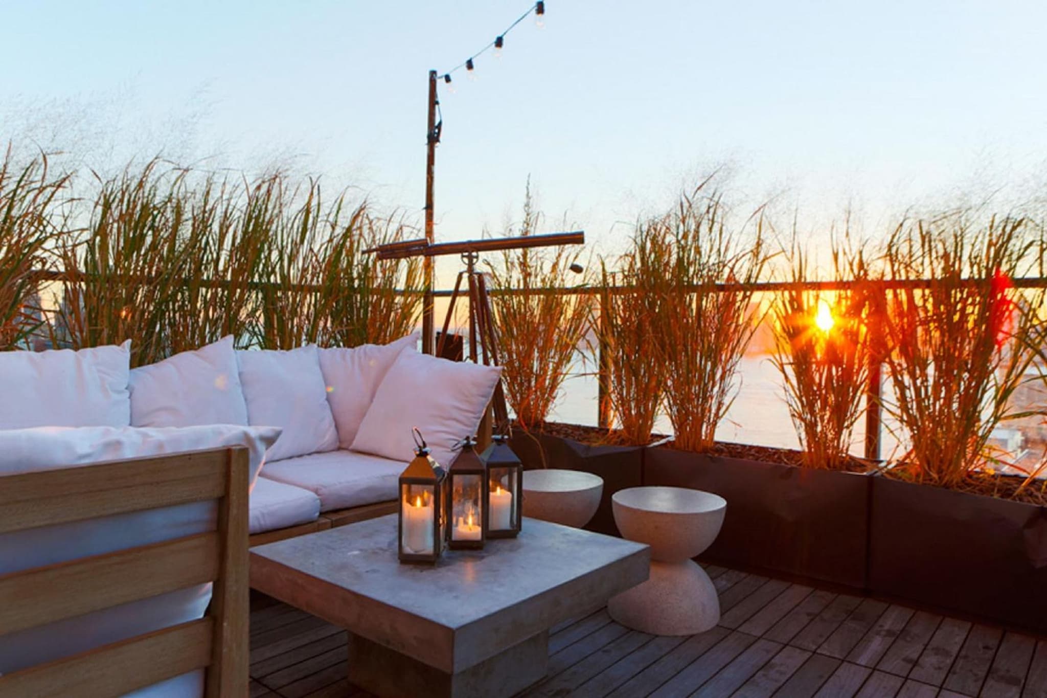 Wooden rooftop deck with white cushioned seating, candle lanterns on a concrete table, tall grasses, and a sunset view.
