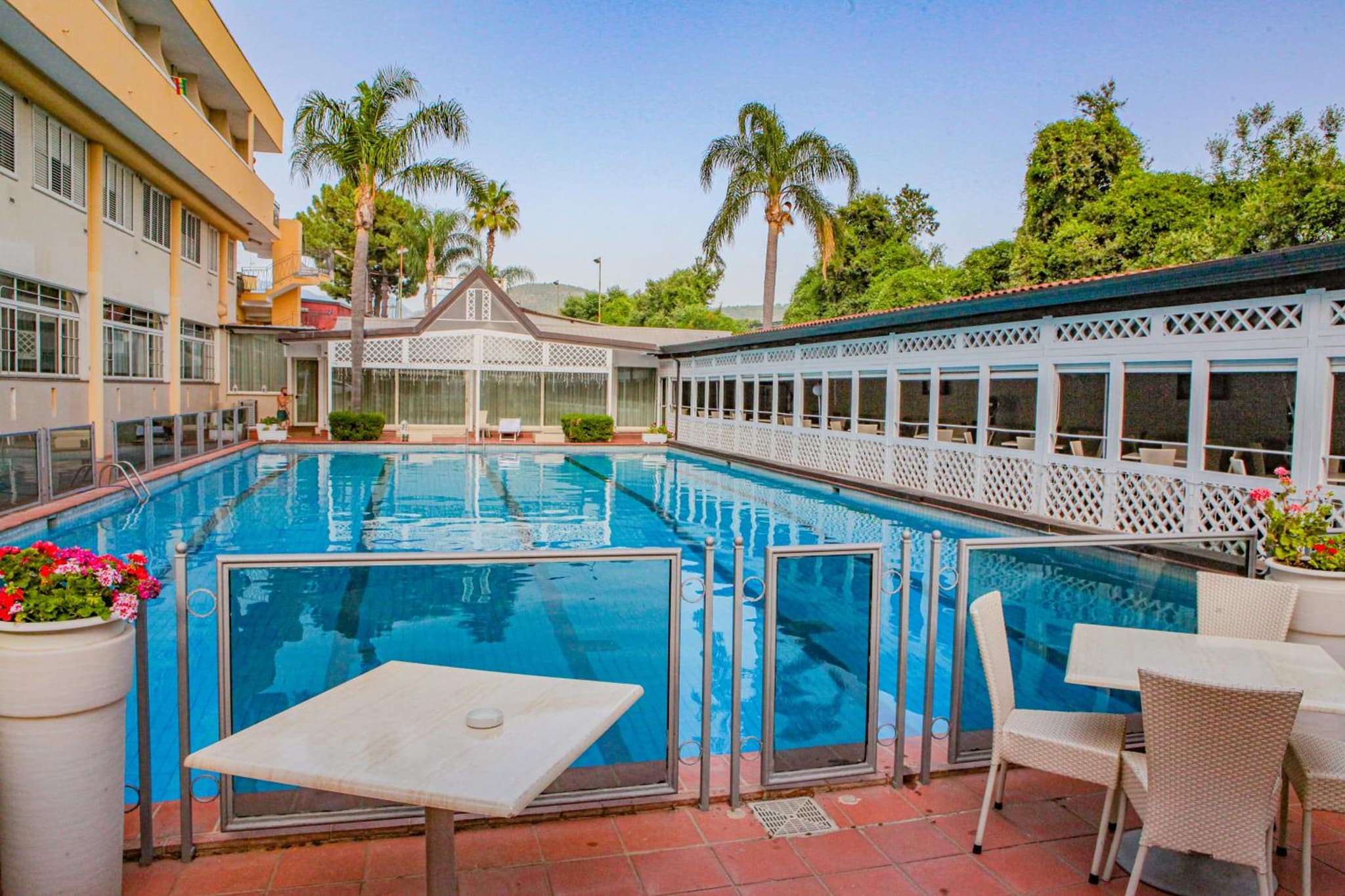 A poolside terrace with outdoor tables and wicker chairs behind glass railings beside the blue pool.