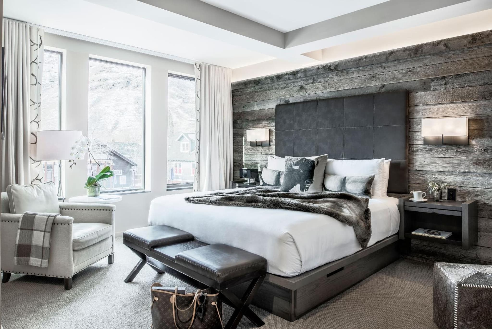 A bedroom with a leather headboard against reclaimed wood paneling, an armchair, and windows facing hills.