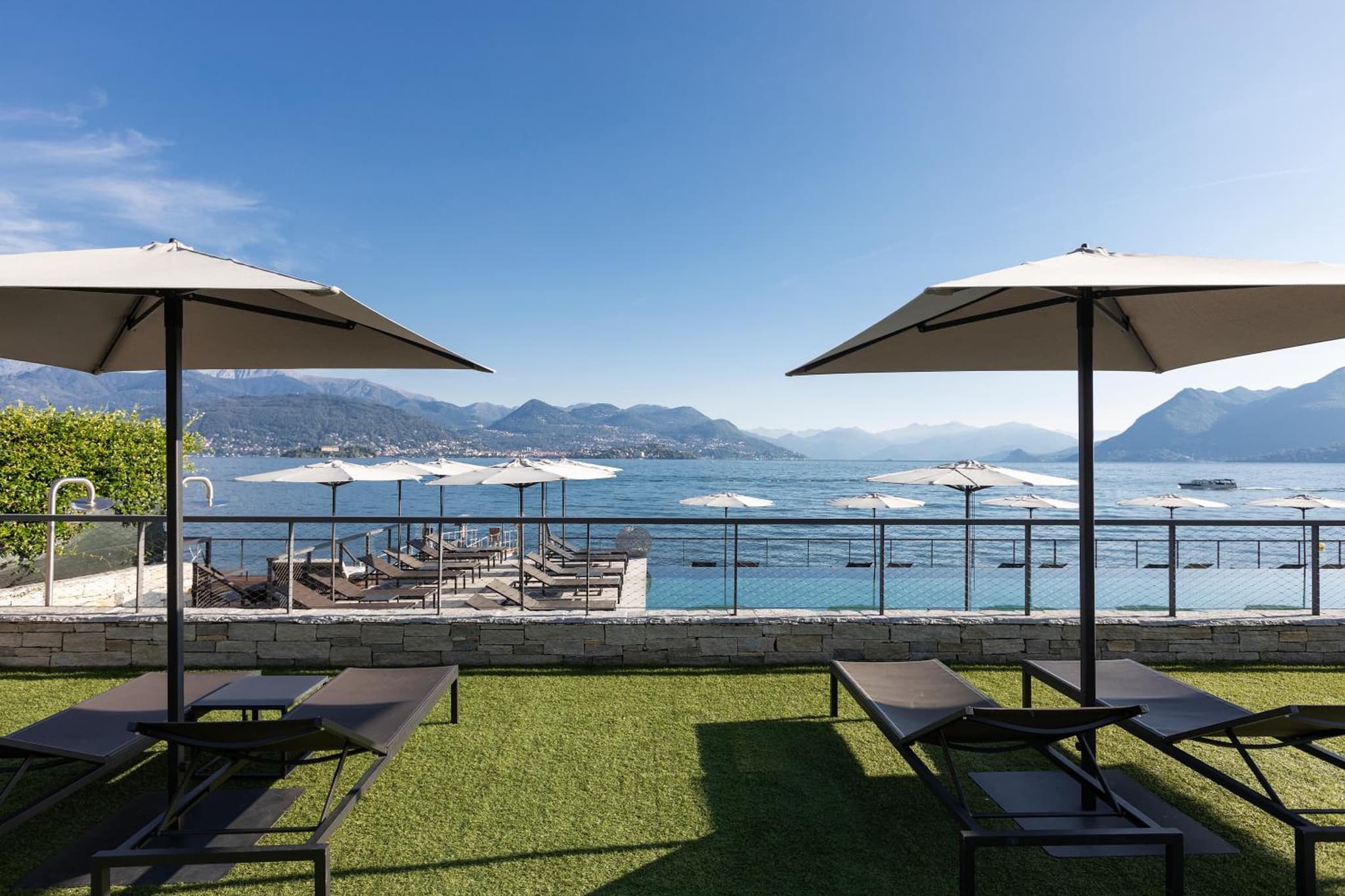 Sun loungers and parasols on a terrace overlooking a pool, open lake waters, and distant mountain peaks.
