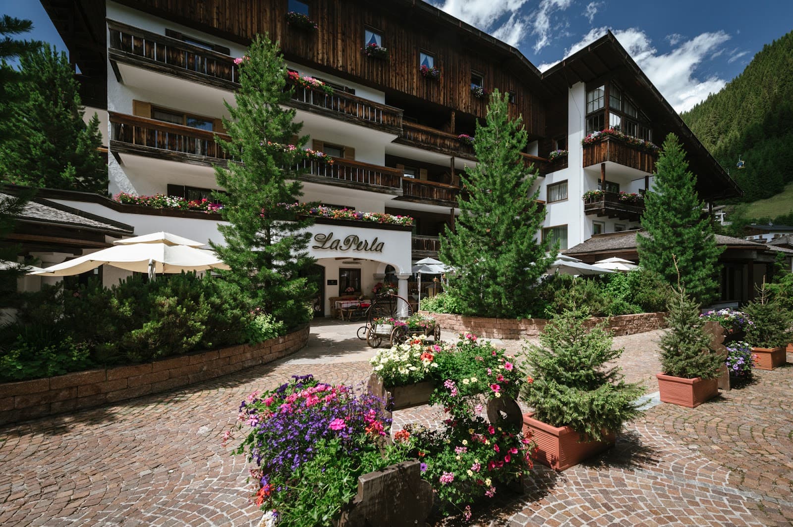 Hotel La Perla facade with flower-lined balconies, evergreen trees, and a cobblestone courtyard beneath a blue sky.