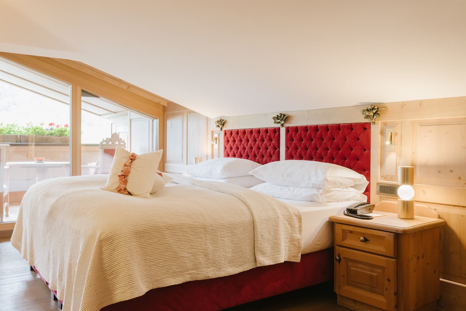 Hotel guest room with a red tufted headboard, cream bedding, light wood panelling, and a glazed balcony door.