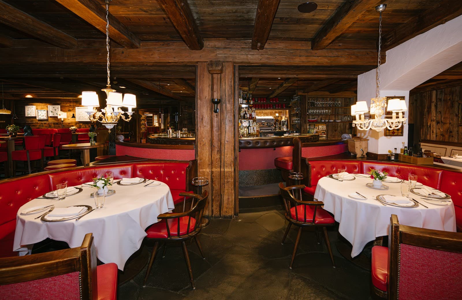 Restaurant interior with red leather booth seating, set round tables with white linen, dark beams, and a central bar.