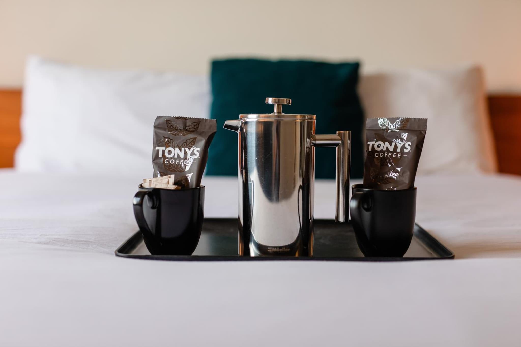 A metal French press and black mugs with coffee packets rest on a tray atop a bed.
