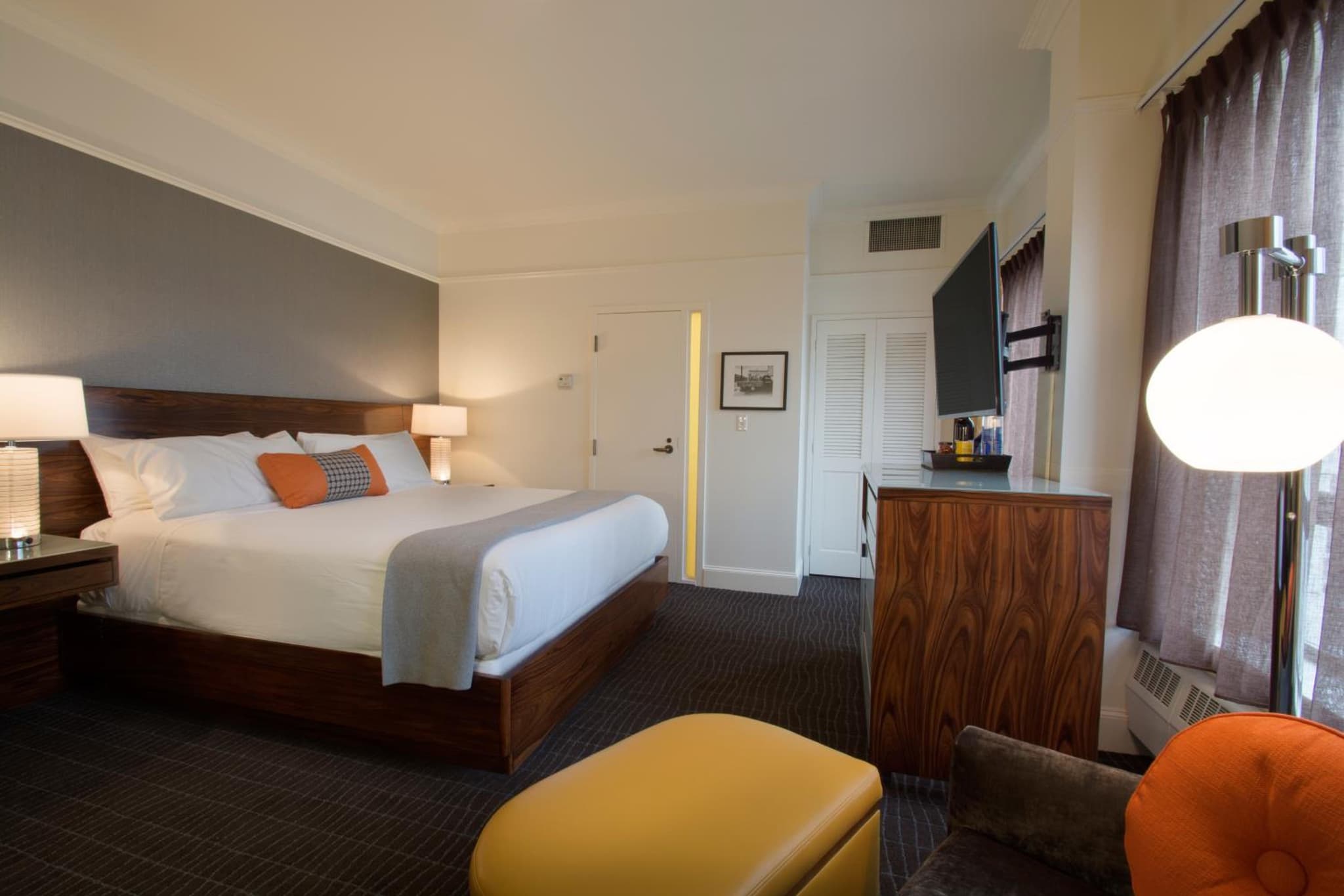 A guest room with a wooden bed frame, gray accent wall, mounted television, and a mustard yellow footstool.