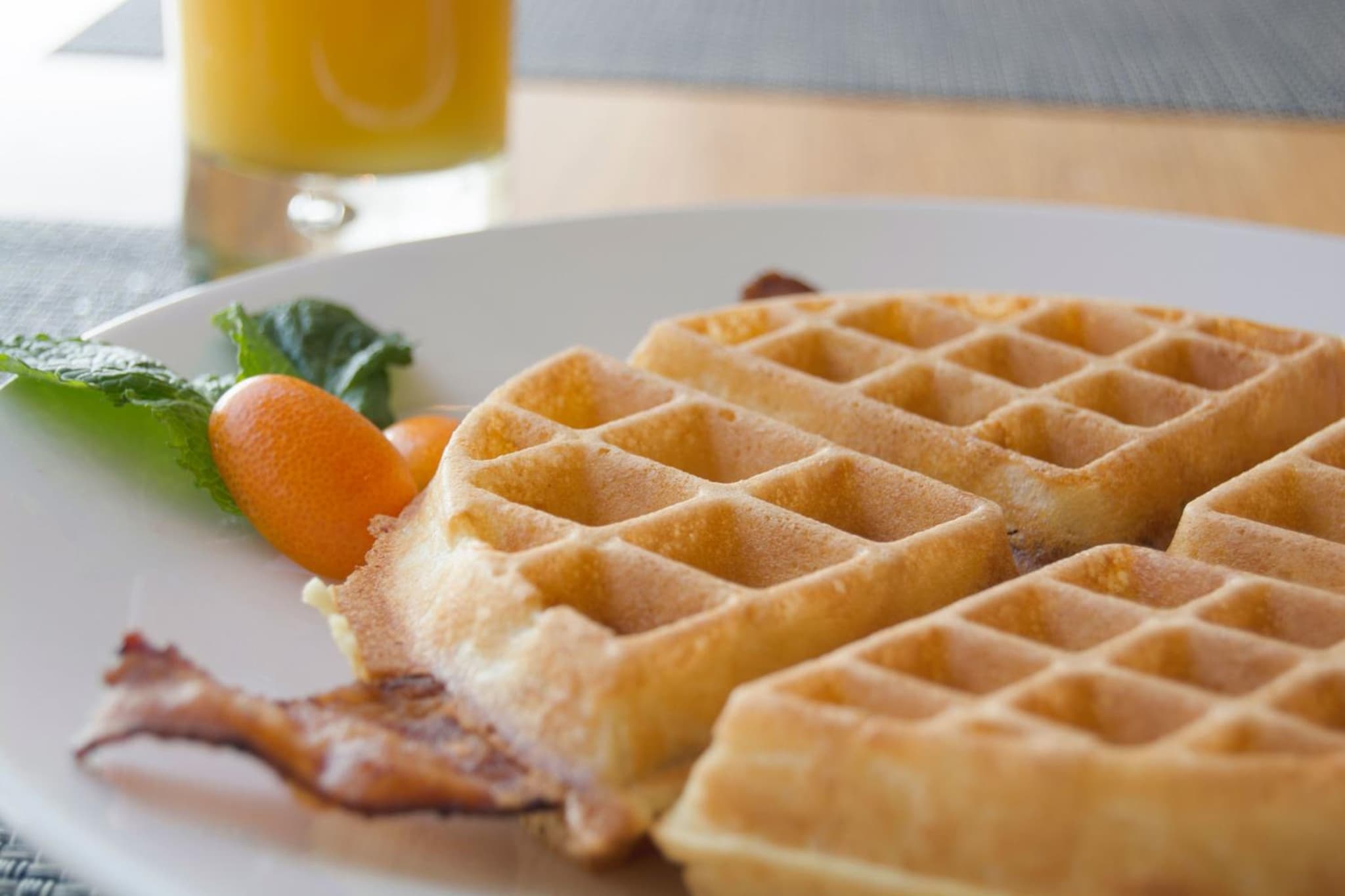 Sliced waffles served with bacon and kumquats on a white plate next to orange juice.