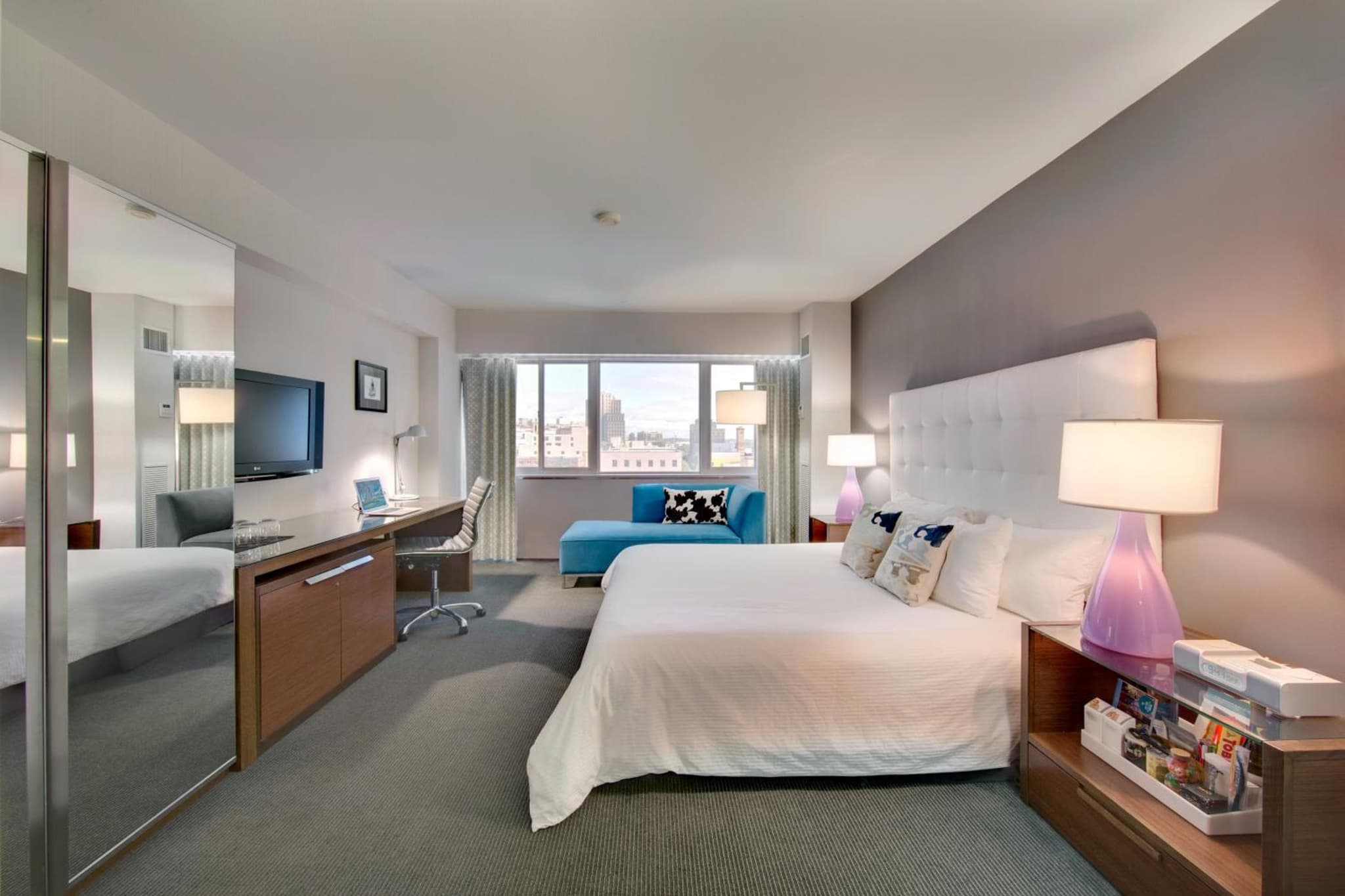 A bedroom with a white tufted headboard, purple bedside lamps, work desk, and a large window.
