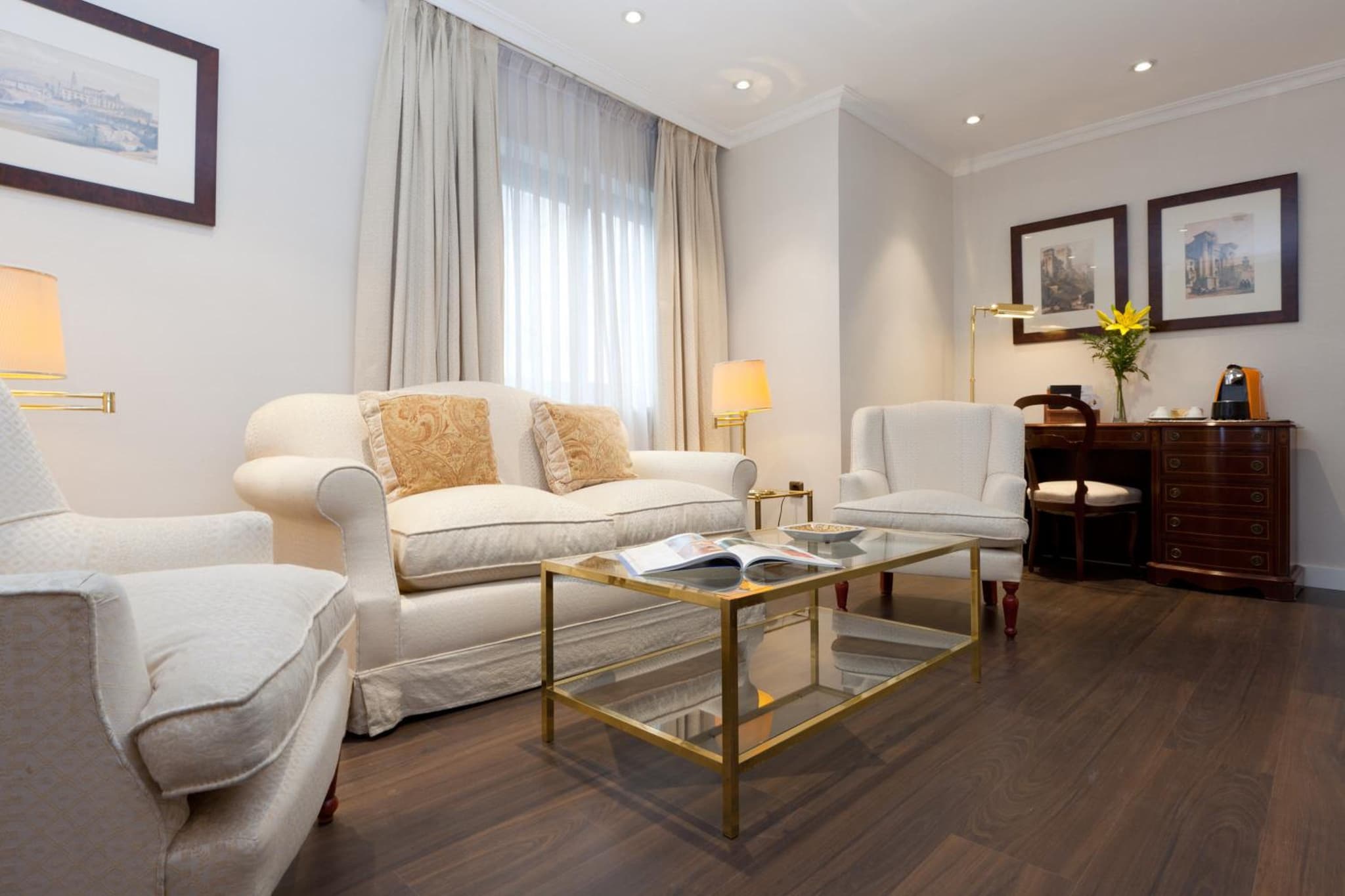 A sitting area with cream armchairs, a fabric sofa, a glass-topped coffee table, and dark wood floors.