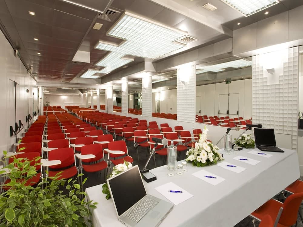 Hotel Savoy conference hall features rows of red chairs facing a draped table with laptops and microphones.