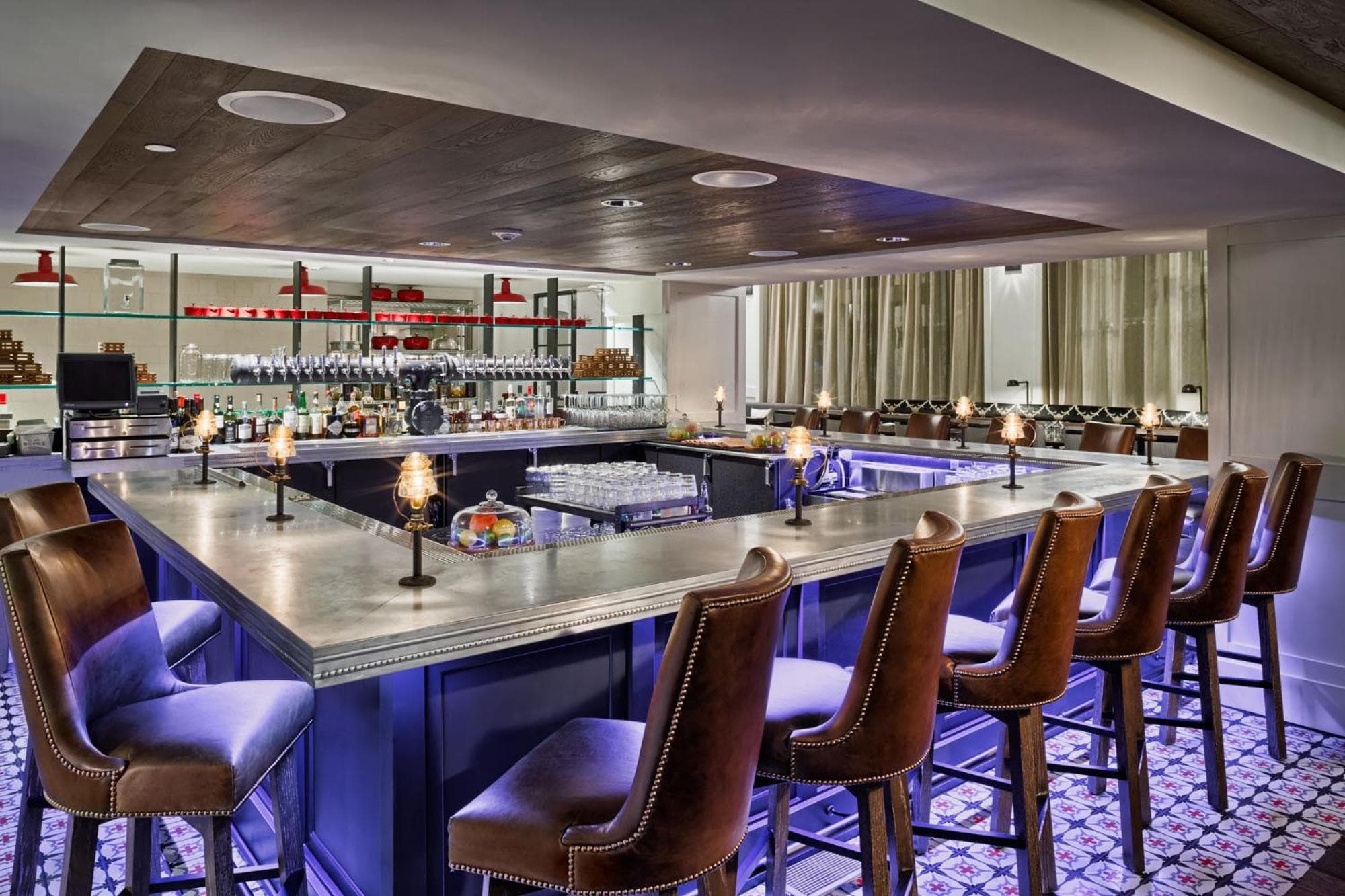 A wrap-around metal bar counter with leather barstools, mood lighting, and patterned floor tile.