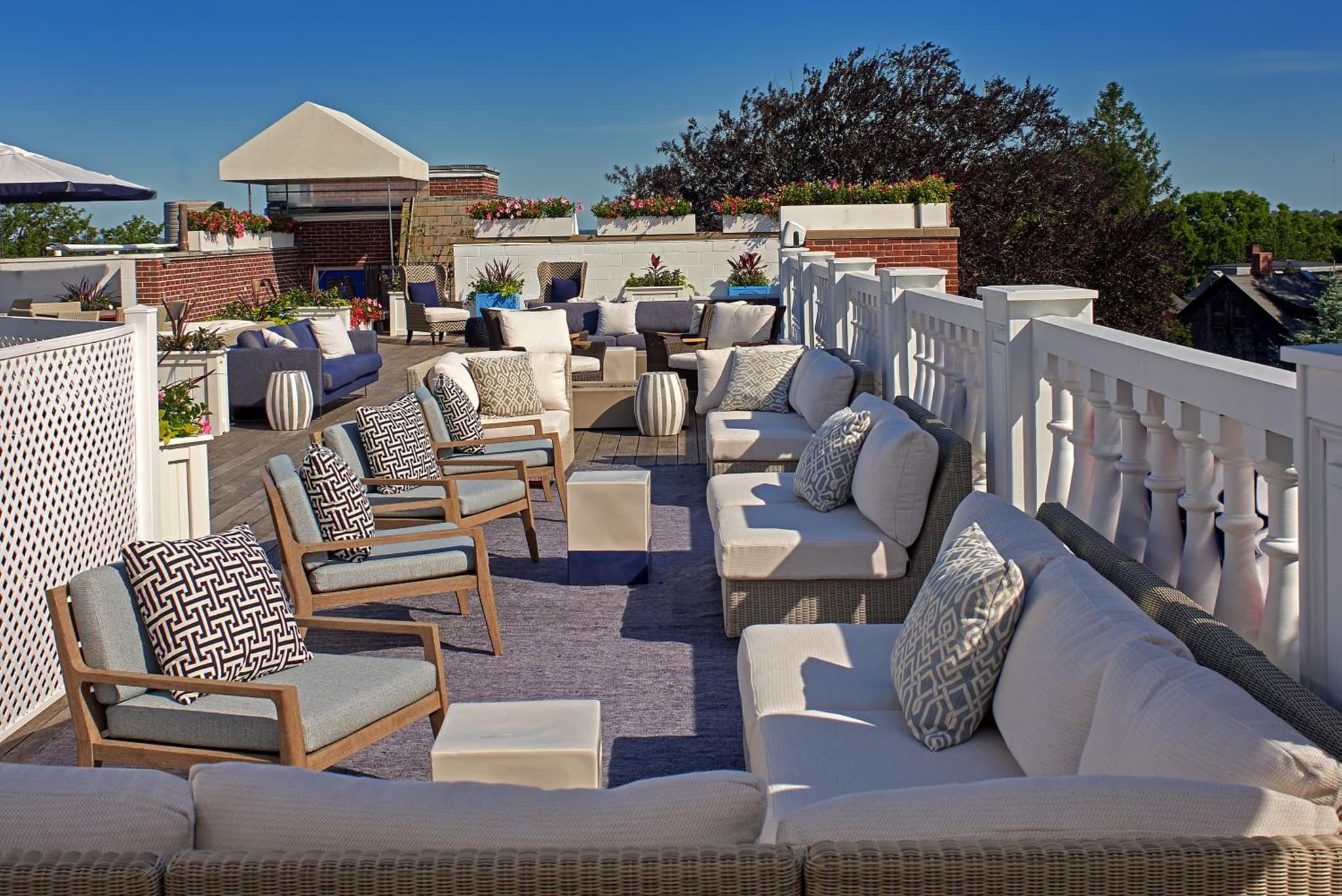 A sunny rooftop terrace furnished with cushioned wicker sofas, armchairs, and planters along a white balustrade.